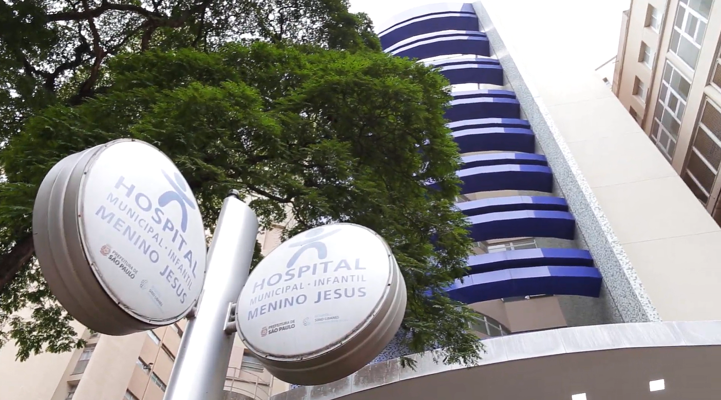 A imagem mostra a fachada do Hospital Municipal Infantil Menino Jesus, localizado em São Paulo. O prédio possui uma arquitetura moderna com varandas onduladas e detalhes em tons de azul. Em primeiro plano, há duas placas redondas destacando o nome do hospital e sua administração pela Prefeitura de São Paulo, em parceria com o Sírio-Libanês. Há também uma grande árvore com folhagem verde, que complementa a paisagem urbana.