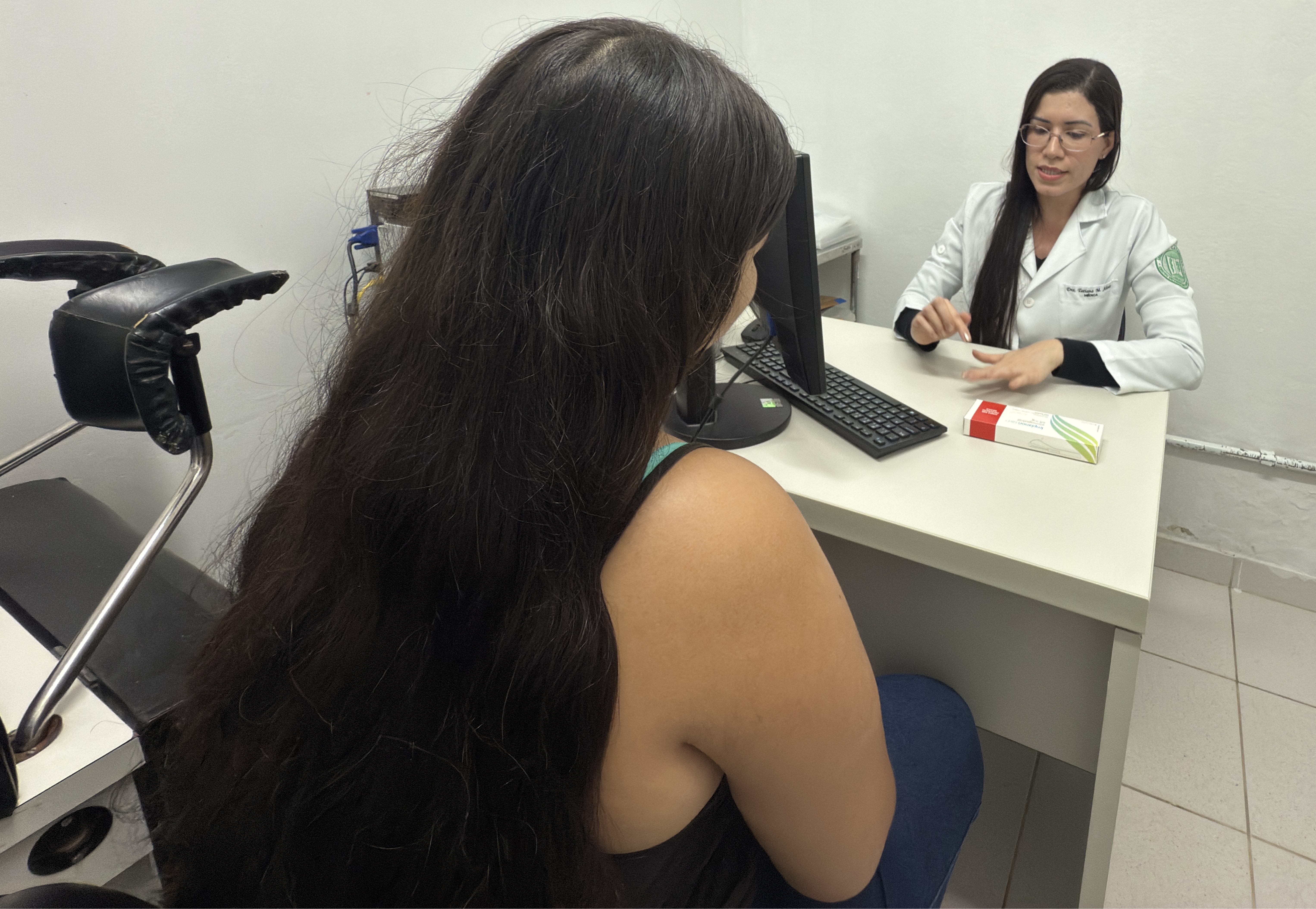 A imagem mostra uma consulta médica em um consultório. Uma profissional de saúde, vestida com um jaleco branco e usando óculos, está sentada atrás de uma mesa, explicando algo para a paciente. A paciente, de costas para a câmera, tem cabelos longos e escuros e veste uma blusa preta. Na mesa, há um computador, um teclado e uma caixa de medicamento. O ambiente é simples, com paredes brancas e poucos móveis. Ao lado da paciente, há uma cadeira ginecológica com apoio para as pernas, sugerindo que a consulta está relacionada à saúde da mulher