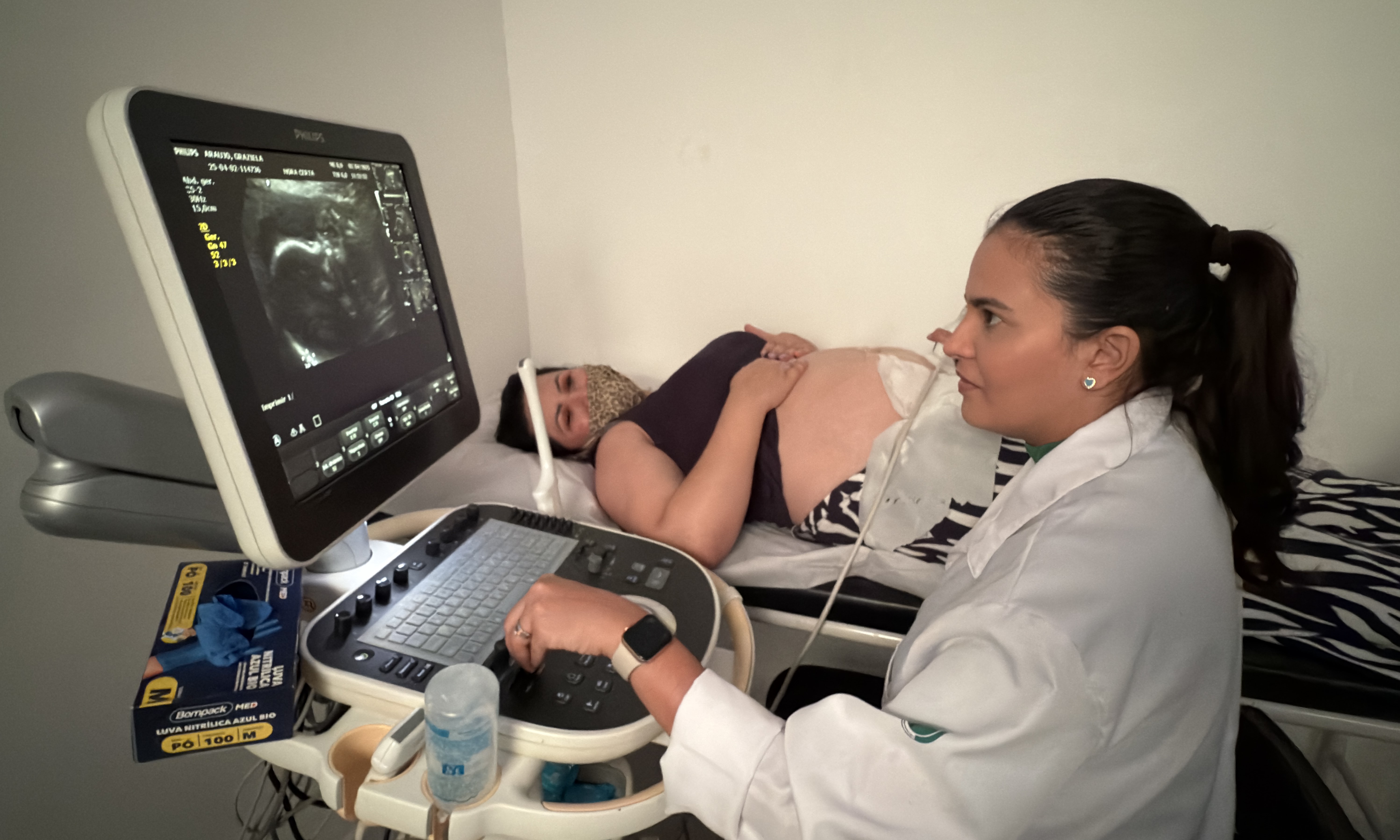 A imagem mostra uma profissional da saúde realizando um exame de ultrassonografia em uma gestante. A mulher grávida está deitada em uma maca, com a barriga exposta e coberta por gel condutor, enquanto observa a tela do equipamento com expressão tranquila. Ela está usando uma máscara facial, o que pode indicar um cuidado com medidas sanitárias. A profissional, que veste jaleco branco e tem o cabelo preso, opera o aparelho de ultrassom, focada nas imagens exibidas no monitor — que mostra claramente o feto dentro do útero. Ao lado do equipamento, há itens de uso clínico como luvas, gel e utensílios médicos. A cena transmite um momento típico de cuidado pré-natal, evidenciando a importância do acompanhamento médico durante a gestação para garantir a saúde da mãe e do bebê.