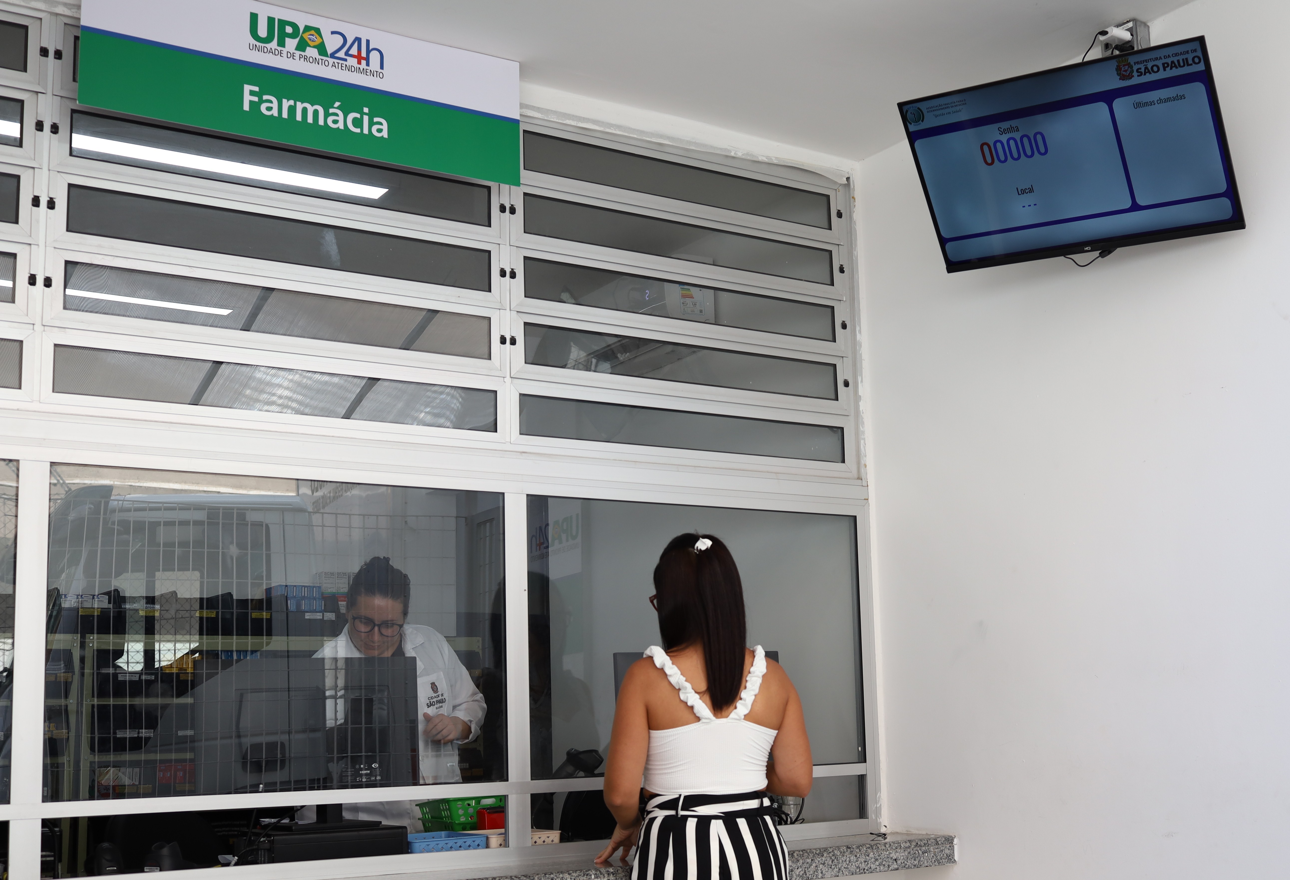 A imagem mostra o balcão de uma farmácia localizada dentro de uma unidade de pronto atendimento (UPA 24h). Uma mulher de costas, vestindo uma blusa branca com detalhes ondulados nas alças e uma saia listrada em preto e branco, está sendo atendida por uma funcionária que veste jaleco branco e está atrás de um vidro, utilizando um computador. Acima do balcão, há uma placa indicando