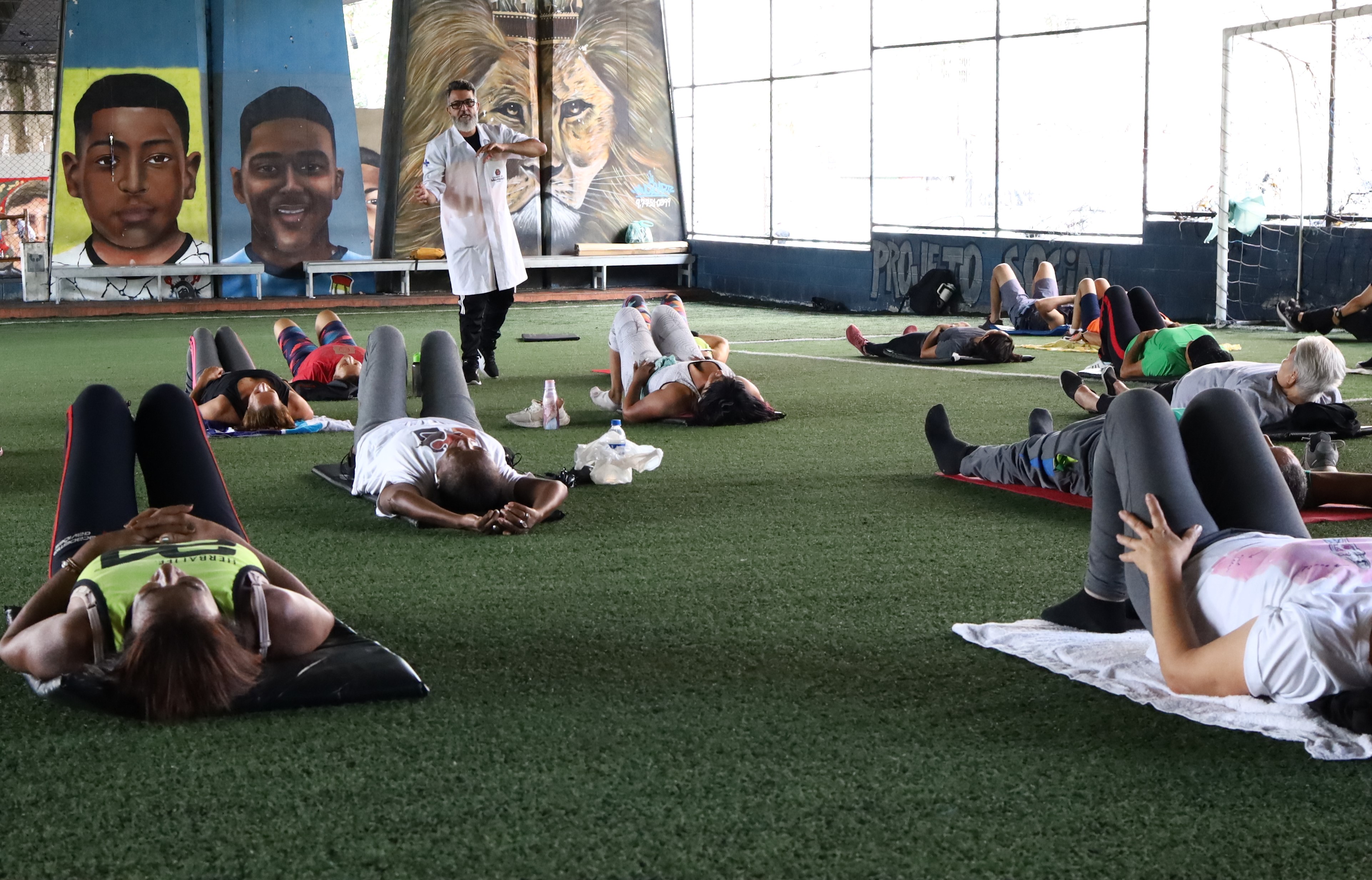 A imagem mostra um grupo de pessoas deitadas de costas em colchonetes, participando de uma atividade física em um espaço coberto, provavelmente uma quadra de esportes. O local possui grama sintética no chão e é decorado com grandes murais nas paredes, incluindo retratos de duas pessoas e a imagem de um leão ao centro. Um instrutor, vestido com jaleco branco, está em pé à frente dos participantes, gesticulando enquanto orienta a atividade. As pessoas parecem estar praticando alongamentos, relaxamento ou alguma prática de atenção corporal, como yoga terapêutica ou exercícios de reabilitação. No fundo, vê-se a estrutura de uma quadra de futsal e parte de uma rede de gol. O ambiente transmite uma sensação de cuidado comunitário e promoção da saúde.