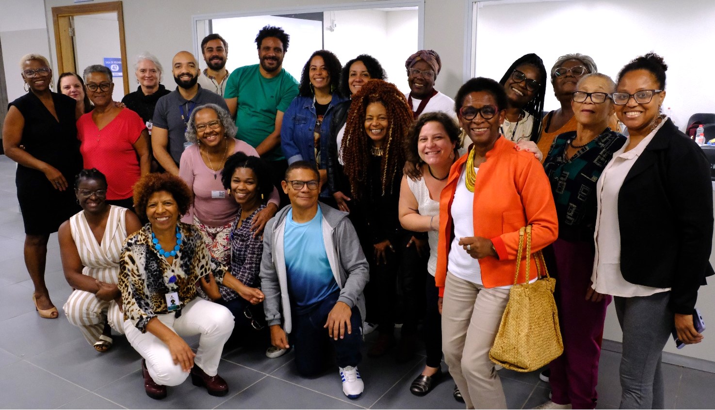 A foto mostra um grupo de cerca de 20 pessoas, homens e mulheres, em sua maioria negras, posando para a foto; elas estão em um corredor, em um ambiente interno