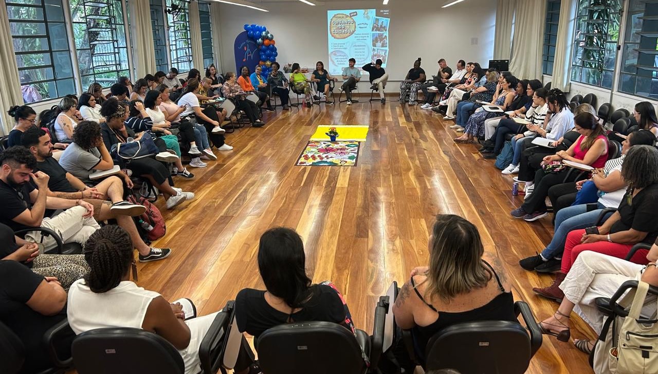 A imagem mostra um grupo grande de pessoas sentadas em cadeiras organizadas em círculo em uma sala ampla com piso de madeira. As janelas grandes nas laterais permitem a entrada de luz natural, e o ambiente é iluminado e acolhedor. No centro do círculo, há um tecido amarelo com uma planta e um tapete colorido, possivelmente usados como parte de uma dinâmica ou atividade de grupo. Na parede ao fundo, um projetor exibe uma apresentação com o título 