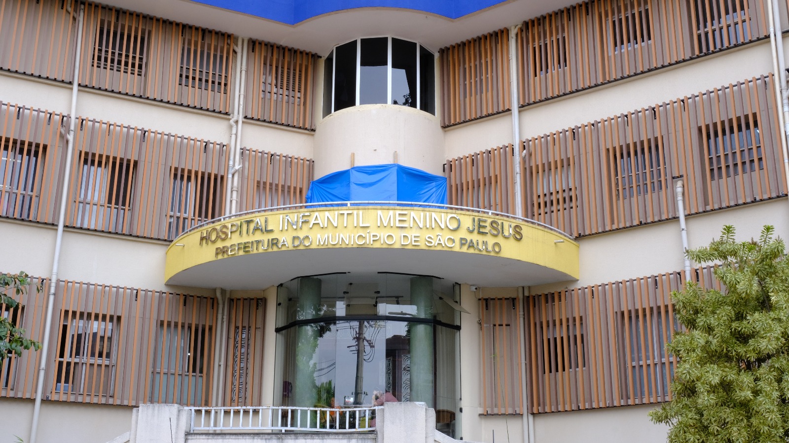 A foto mostra a fachada do Hospital Infantil Menino Jesus, que pertence à Prefeitura do Município de São Paulo. A entrada possui um letreiro em letras douradas com o nome do hospital, e o prédio tem várias janelas protegidas por estruturas verticais de madeira ou metal na cor marrom claro. Acima da entrada há uma estrutura curva, com uma área azul cobrindo parte superior. Há algumas plantas à direita da imagem.