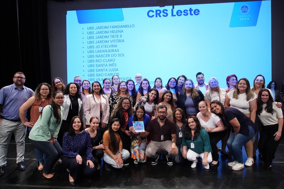 A foto mostra um grupo grande de pessoas reunidas no palco de um auditório. Elas posam sorrindo em frente a um telão que exibe o texto 