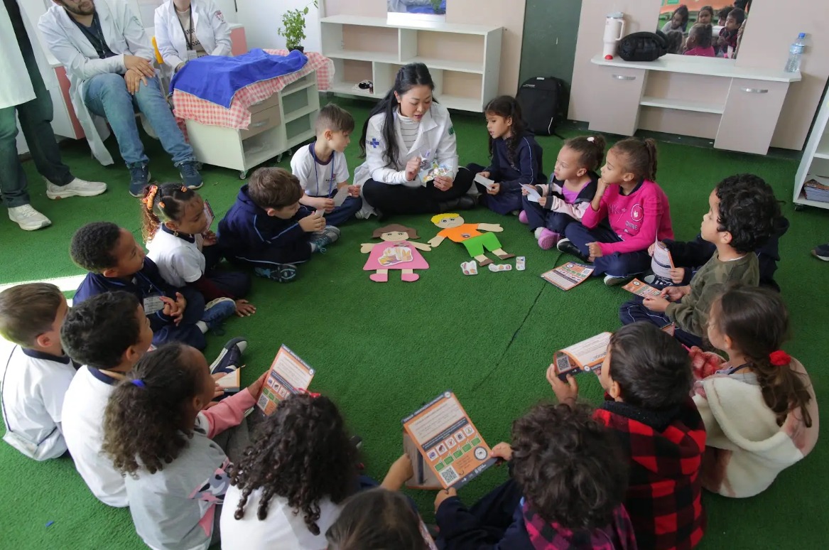 A imagem mostra uma atividade educacional em uma sala, onde um grupo de crianças está sentado em círculo sobre um tapete verde. Elas estão acompanhadas por adultos, profissionais de saúde vestidos com jalecos brancos. No centro do círculo, há recortes de figuras humanas em papel, que parecem estar sendo usados para uma demonstração ou explicação. As crianças estão atentas e seguram materiais impressos em mãos, que podem ser folhetos ou cartilhas relacionadas ao tema da atividade. A expressão das crianças demonstra envolvimento com o momento, sugerindo que a atividade é interativa e educativa. Ao fundo, há móveis simples e organizados, como estantes e mesas, reforçando o ambiente acolhedor e funcional para aprendizado.