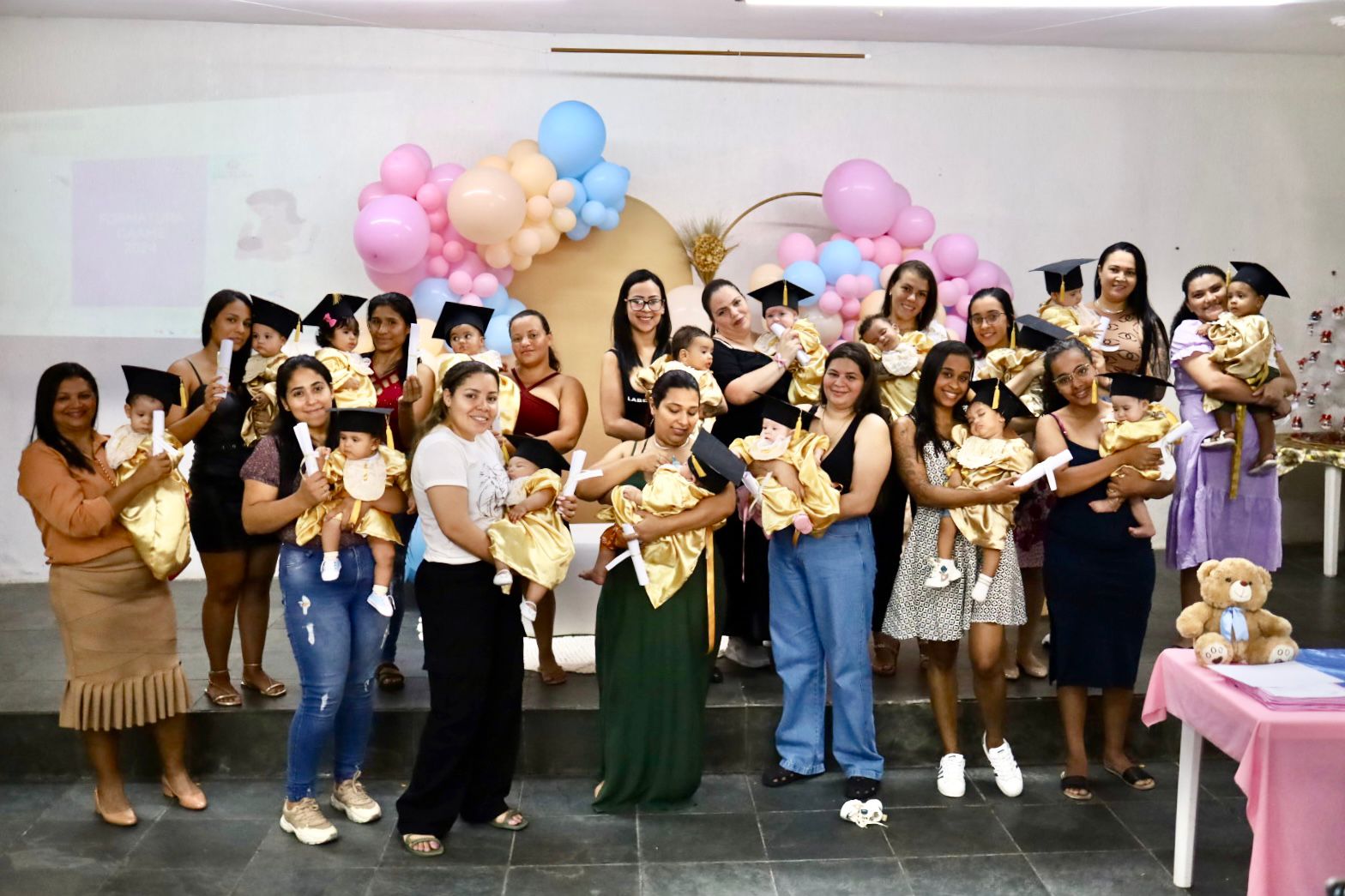 A imagem mostra um grupo de mulheres segurando bebês vestidos com roupas douradas e chapéus de formatura. As mulheres estão sorrindo e algumas seguram diplomas simbólicos, sugerindo uma celebração ou formatura. O fundo está decorado com balões em tons de rosa, azul e dourado, criando um ambiente festivo e acolhedor