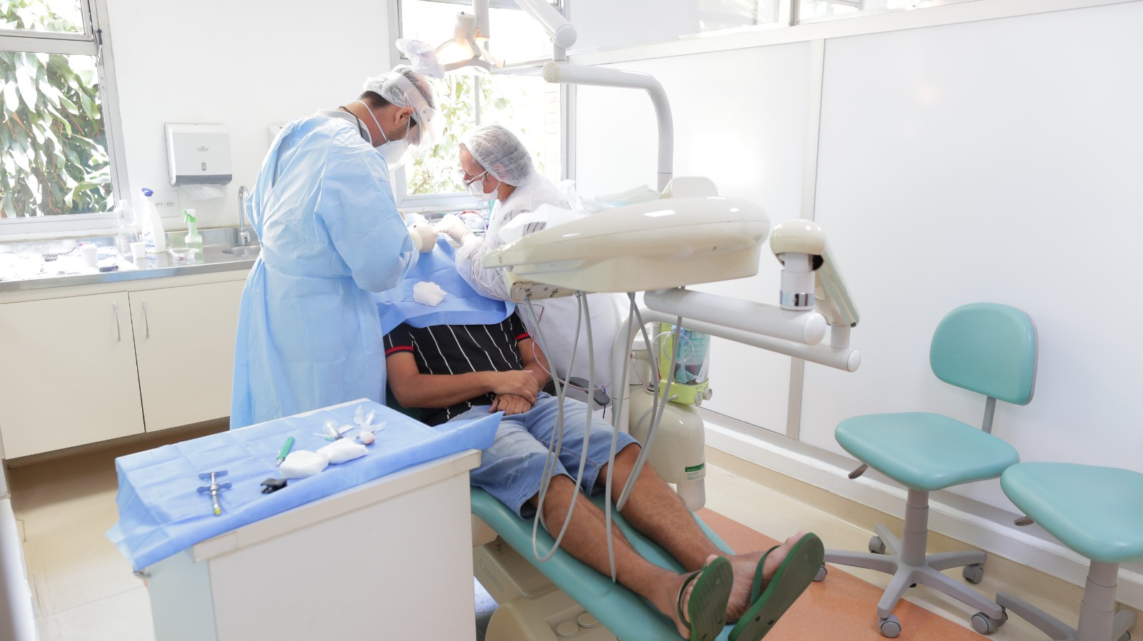 A imagem mostra uma consulta odontológica em andamento. O paciente está deitado em uma cadeira odontológica, vestindo bermuda jeans e camiseta listrada, com os pés calçados em chinelos. Dois profissionais de saúde, usando jalecos, toucas, máscaras e luvas, trabalham no atendimento do paciente. Um deles está à esquerda, de costas, e o outro está ao lado do paciente, mexendo no procedimento.  O consultório apresenta um ambiente limpo e bem iluminado, com paredes brancas e uma ampla janela, que permite a entrada de luz natural. Há equipamentos odontológicos tradicionais, como a cadeira, um refletor e instrumentos cirúrgicos organizados sobre uma bandeja coberta por um tecido azul.