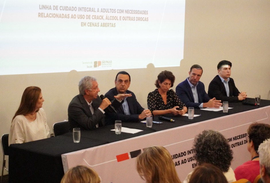 Mesa de abertura do evento com autoridades: são duas mulheres e quatro homens, há seis copos com água e quatro folhas de papel, um deles fala e gesticula ao microfone enquanto os demais observam atentamente