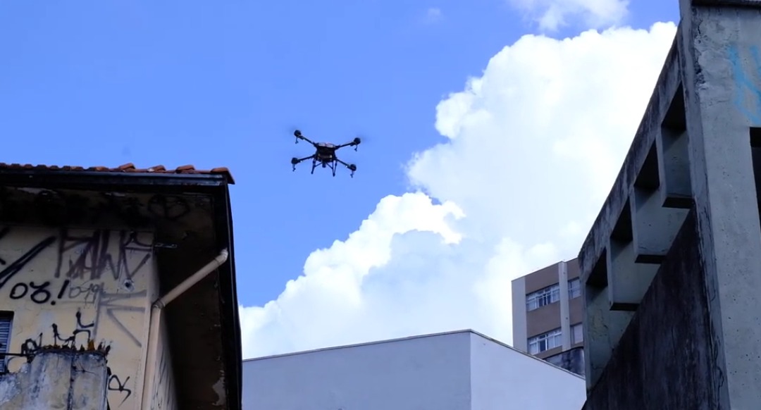 A imagem mostra um drone voando em meio a prédios em uma área urbana durante o dia. O céu está azul com nuvens brancas ao fundo. À esquerda, há um edifício antigo com grafites e sinais de desgaste, como rachaduras e sujeira. À direita, um prédio com arquitetura mais moderna, mas igualmente desgastado, aparece parcialmente