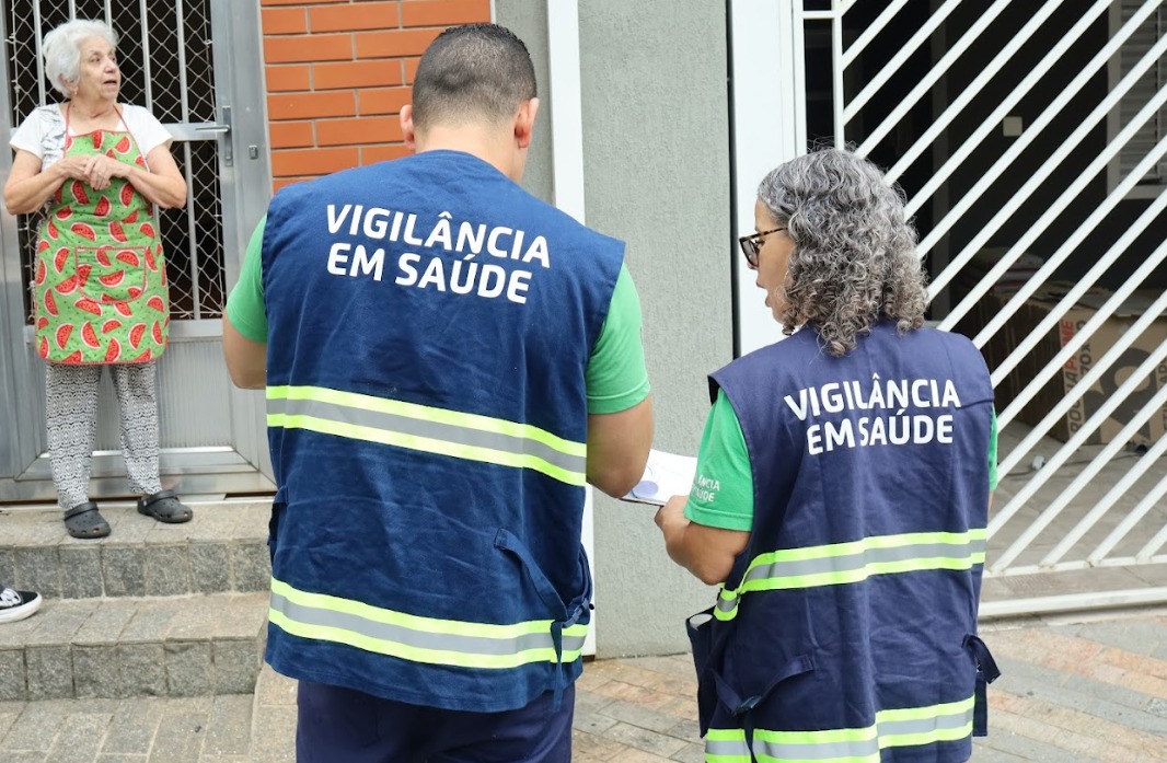 A imagem mostra dois agentes da Vigilância em Saúde realizando uma visita domiciliar. Eles estão de costas, vestindo coletes azuis com faixas refletivas e a inscrição 