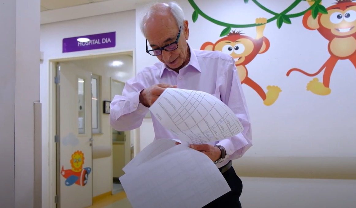 A imagem mostra um homem idoso, de cabelos brancos, usando óculos e vestindo uma camisa social clara. Ele está segurando e analisando folhas de papel com tabelas ou informações escritas, aparentando estar em um ambiente hospitalar. O local possui uma decoração infantil, com desenhos coloridos de macacos pendurados em cipós nas paredes, e um adesivo de um leão em um avião próximo à porta. Acima dessa porta, há uma placa roxa com a inscrição