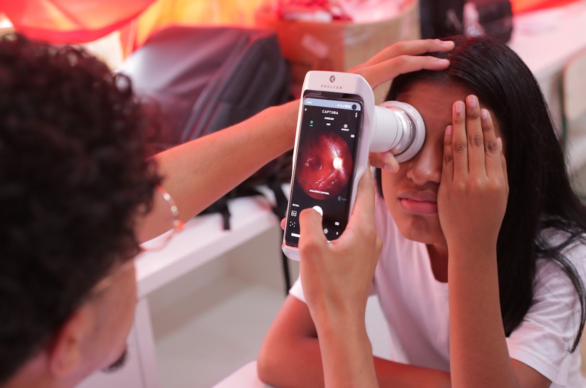 A imagem mostra uma cena em que uma pessoa está realizando um exame oftalmológico em outra pessoa. O examinador utiliza um dispositivo eletrônico moderno conectado a um smartphone para capturar imagens do interior do olho, aparentemente do fundo ocular. A pessoa que está sendo examinada cobre um dos olhos com a mão, enquanto o outro olho é analisado. Ao fundo, há mochilas e uma luz avermelhada difusa que ilumina parcialmente o ambiente