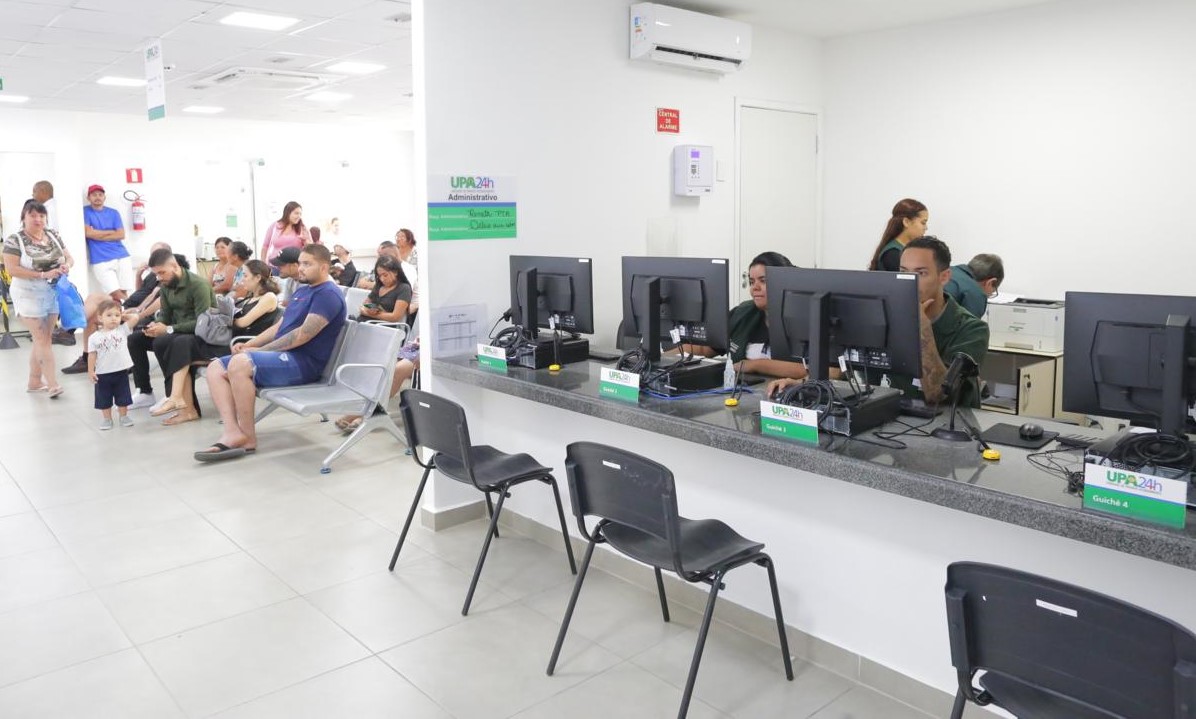 A imagem mostra a recepção de uma Unidade de Pronto Atendimento (UPA) 24h, um espaço de atendimento médico emergencial do sistema público de saúde. Em primeiro plano, há um balcão de atendimento com quatro guichês, identificados com placas que indicam