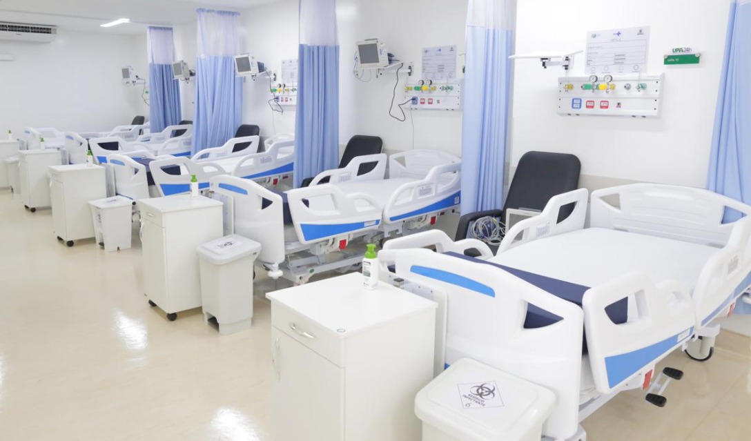 A imagem mostra uma ala hospitalar moderna e bem equipada. Há várias camas hospitalares ajustáveis, com cabeceiras eleváveis, organizadas em fileiras. Cada cama possui equipamentos médicos ao lado, como monitores de sinais vitais e pontos de acesso para oxigênio e outros insumos, fixados na parede. Entre as camas, há cortinas azuis para oferecer privacidade aos pacientes. Também estão presentes armários e recipientes de descarte hospitalar, todos dispostos de forma ordenada. O ambiente é limpo, claro e iluminado, com pisos brilhantes e paredes brancas, refletindo um alto padrão de higiene e organização