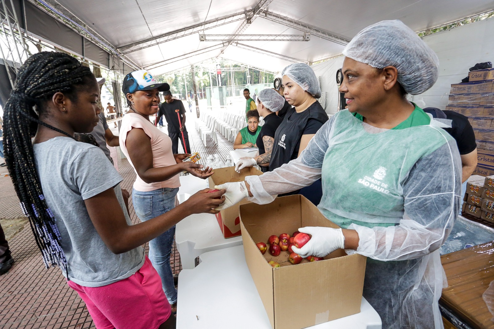 A imagem mostra uma cena de distribuição de alimentos em um espaço coberto. No primeiro plano, uma mulher vestindo avental e touca protetora está entregando frutas a uma jovem. A jovem está recebendo a fruta com as mãos estendidas, demonstrando interação direta. Ao fundo, outras pessoas estão organizando a distribuição de alimentos ou aguardando atendimento. Há caixas de papelão com frutas, como maçãs, no local. O ambiente é iluminado, com uma estrutura de tendas, e o clima parece ser de cooperação e solidariedade. No uniforme da mulher, é possível identificar o emblema ou logo da