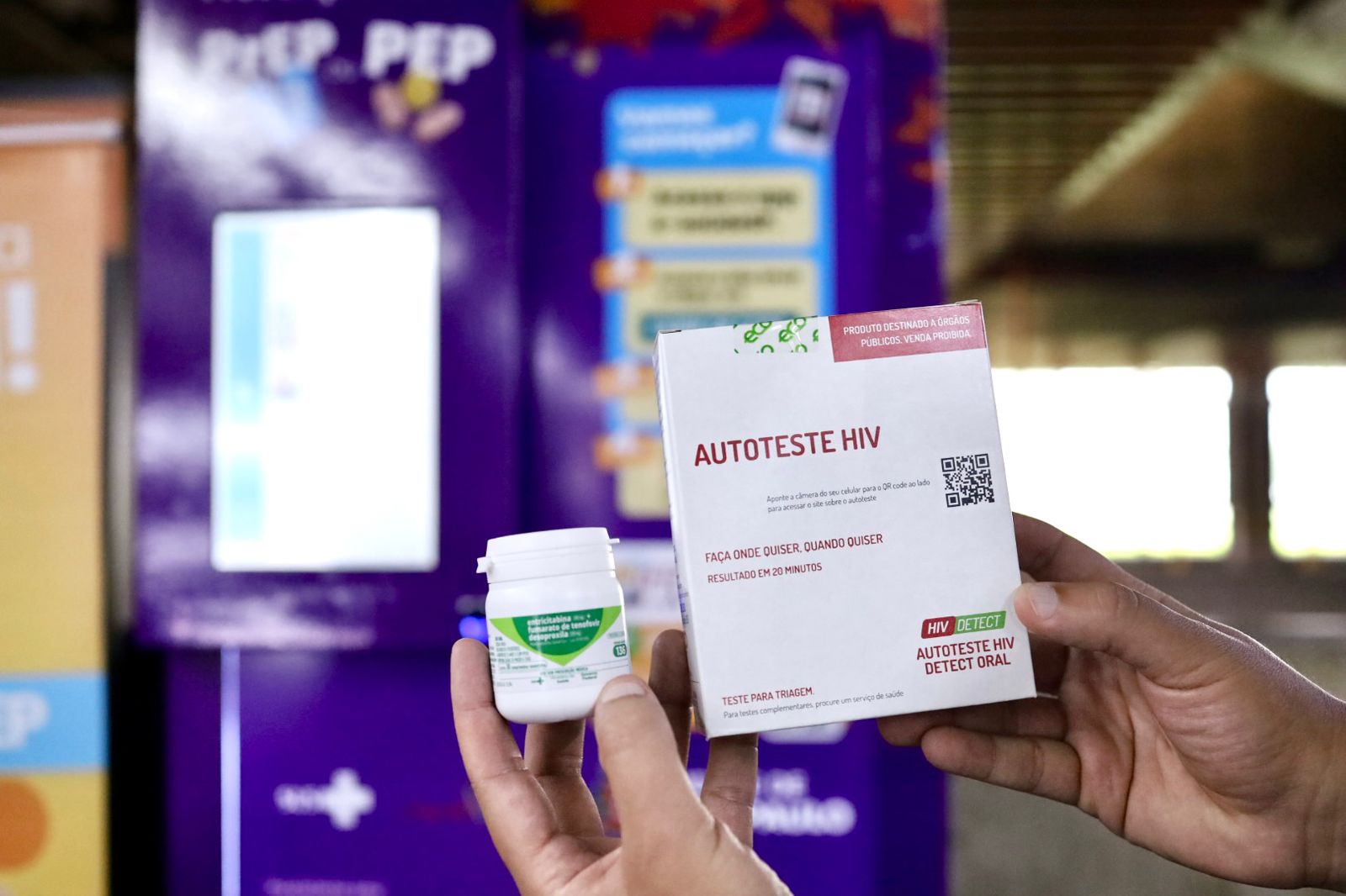 A imagem mostra duas mãos segurando produtos relacionados à prevenção e diagnóstico do HIV. Na mão esquerda, está um frasco branco com tampa de rosca, possivelmente contendo medicamentos (como antirretrovirais ou PrEP). Na mão direita, está uma caixa de autoteste HIV com a inscrição: