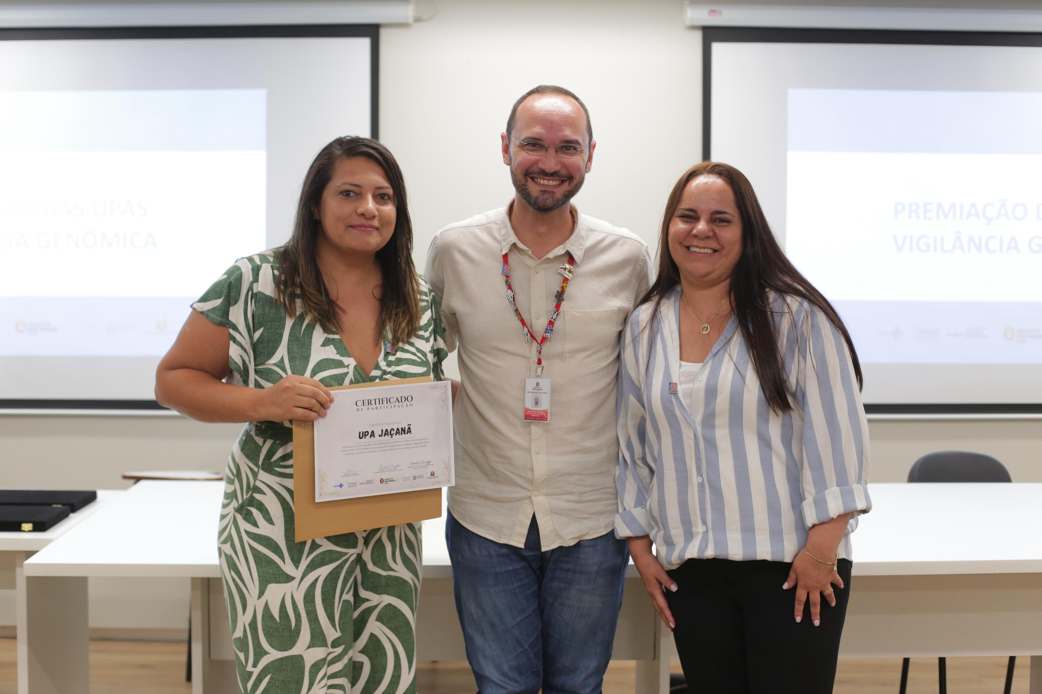 A imagem mostra três pessoas sorrindo e posando para uma foto em um evento de premiação. A mulher à esquerda está segurando um certificado que indica a participação da 