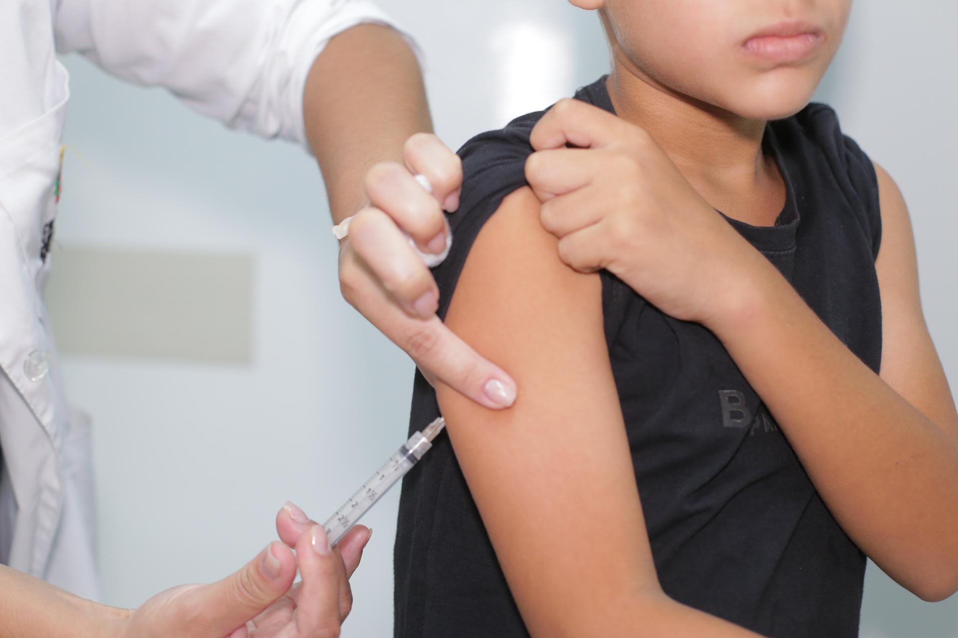 A imagem mostra um profissional de saúde aplicando uma vacina no braço de uma criança. A criança veste uma camiseta preta e levanta a manga para expor o braço, enquanto segura a própria pele com a outra mão. O profissional de saúde, que usa um jaleco branco, segura a seringa e aponta para o local da aplicação. O fundo é um ambiente clínico, com paredes brancas e um clima típico de unidade de saúde. A cena destaca a importância da vacinação infantil para a prevenção de doenças