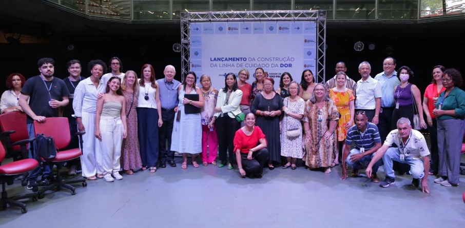A imagem mostra um grupo diversificado de pessoas reunidas para um evento, posando para uma foto em frente a um painel que contém a inscrição