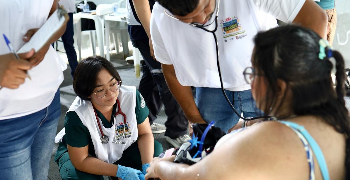 A imagem mostra uma equipe de profissionais de saúde realizando um atendimento. No centro, uma profissional de jaleco branco e roupa verde, com um estetoscópio no pescoço, está ajoelhada e observando atentamente a paciente, demonstrando atenção e cuidado. Ao seu lado, outro profissional de saúde, vestindo uma camiseta branca com um crachá e um estetoscópio, está medindo a pressão arterial de uma mulher de cabelos escuros, que veste uma blusa de alças azuis. No canto esquerdo da imagem, uma pessoa segura uma prancheta e parece anotar informações. O fundo indica um ambiente movimentado, com outras pessoas e cadeiras brancas, sugerindo uma ação de saúde comunitária, possivelmente em um evento público ou mutirão de atendimento