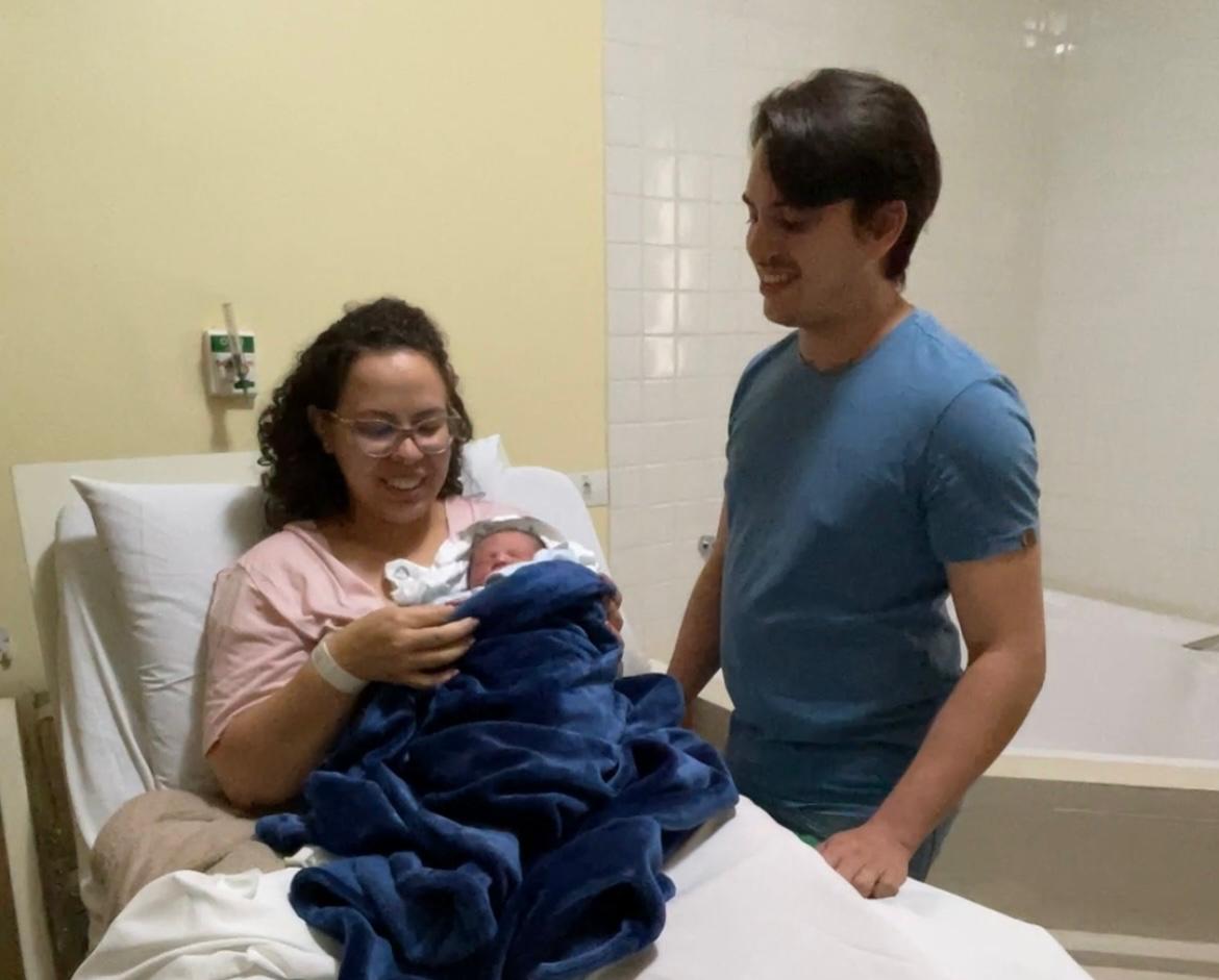 A imagem mostra uma cena emocionante em um quarto de hospital. Uma mulher está sentada em uma cama hospitalar, segurando um recém-nascido enrolado em um cobertor azul. Ela sorri enquanto olha para o bebê. Ao lado dela, um homem de camisa azul, possivelmente o pai da criança, também sorri e observa a mãe e o bebê com carinho. O ambiente tem paredes de tom claro e equipamentos hospitalares ao fundo, sugerindo que o nascimento ocorreu recentemente. A expressão dos pais transmite felicidade e ternura.