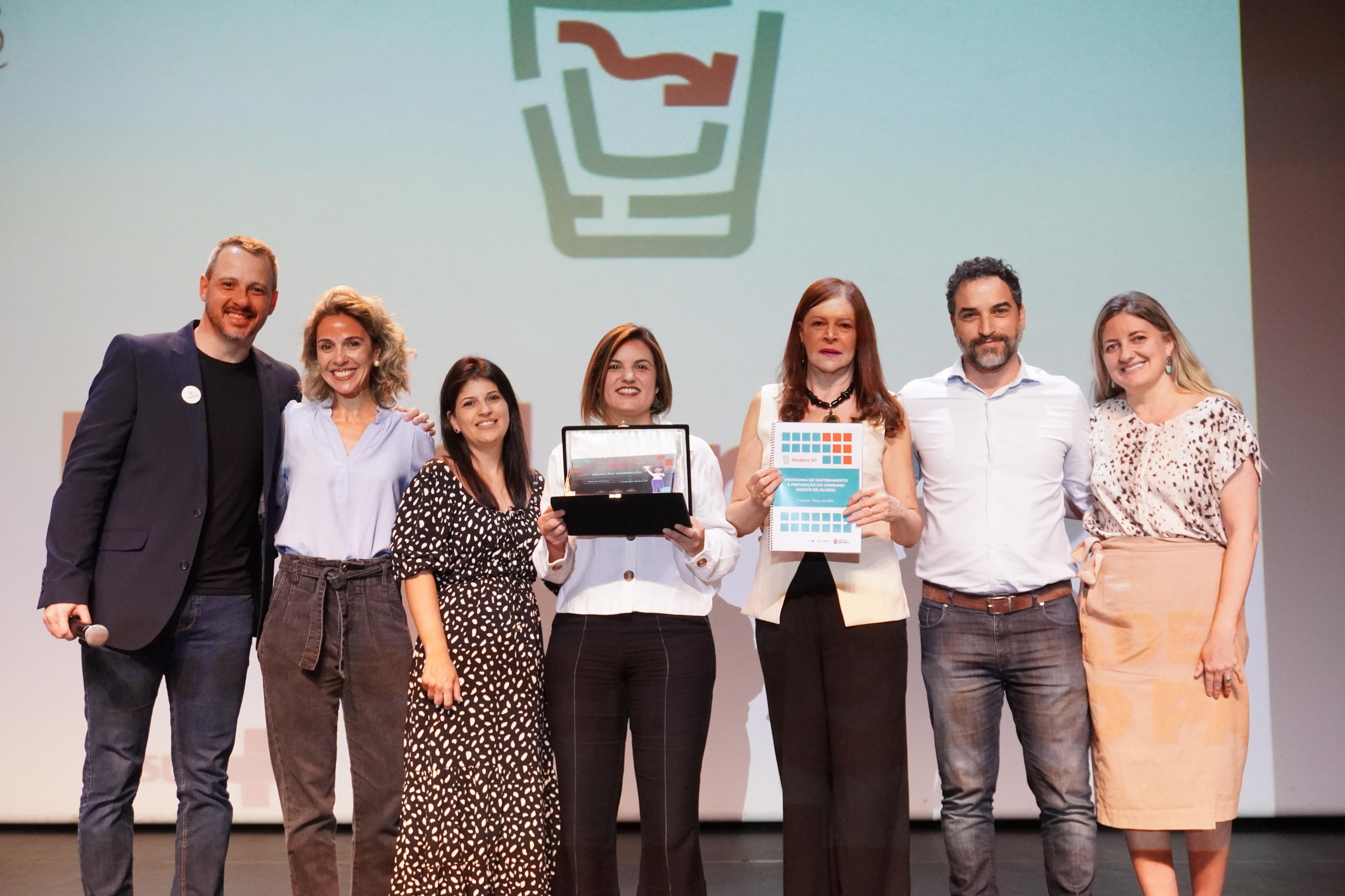 A foto mostra um grupo de sete pessoas posando em um palco, sorrindo e comemorando uma premiação ou reconhecimento. No centro, uma mulher segura um troféu em uma caixa preta, enquanto outra mulher, ao lado, segura um documento com uma capa colorida, que parece ser um certificado ou relatório. Todos estão vestidos de maneira formal ou casual elegante. O fundo da imagem exibe um telão com um logotipo estilizado que lembra um copo ou símbolo gráfico. O clima da foto é de celebração e conquista.