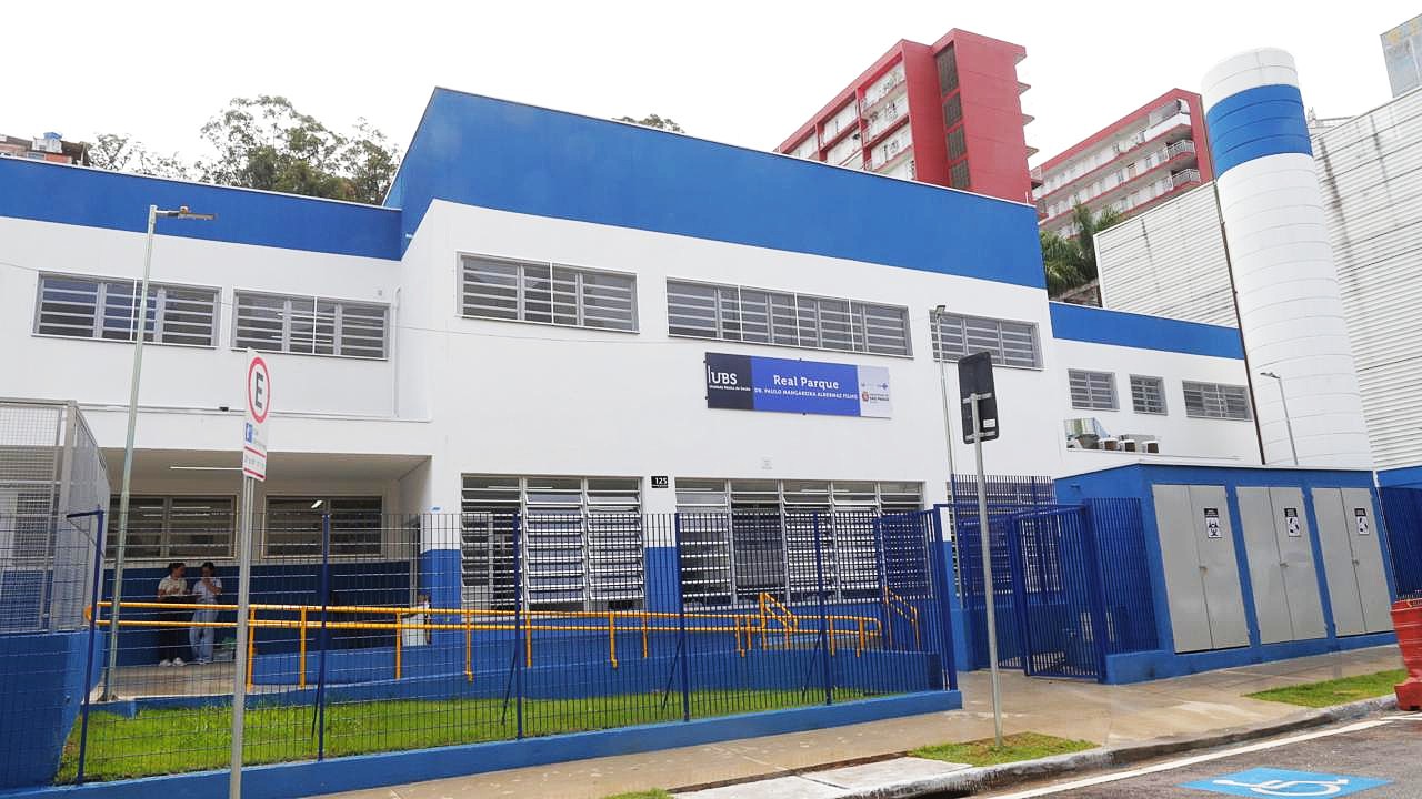 A imagem mostra a fachada de uma UBS (Unidade Básica de Saúde) localizada no bairro Real Parque, com um design moderno. O prédio tem uma combinação de cores azul e branco, e é de dois andares, com janelas grandes. Em frente, há uma cerca de metal azul, com uma rampa de acesso amarelo. No canto inferior esquerdo, é possível ver duas pessoas sentadas em uma área coberta. Um cartaz visível na fachada indica o nome da unidade de saúde. O ambiente ao redor está bem cuidado, com calçadas e sinalização de acessibilidade.