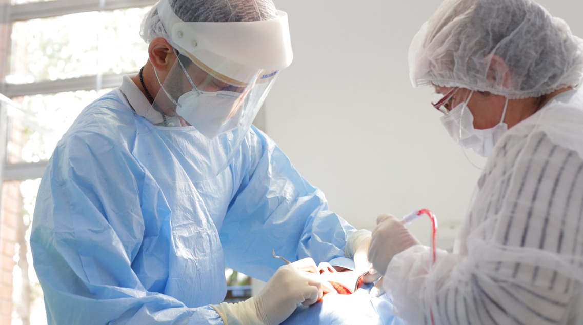 A imagem mostra dois profissionais de saúde em um ambiente clínico, provavelmente realizando um procedimento cirúrgico. Ambos estão usando roupas de proteção, como aventais azuis, luvas e máscaras cirúrgicas, além de protetores faciais. O profissional à esquerda está com um capacete protetor e está focado em um procedimento, enquanto o outro, à direita, também usa um protetor na cabeça e está assistindo ou ajudando no processo.