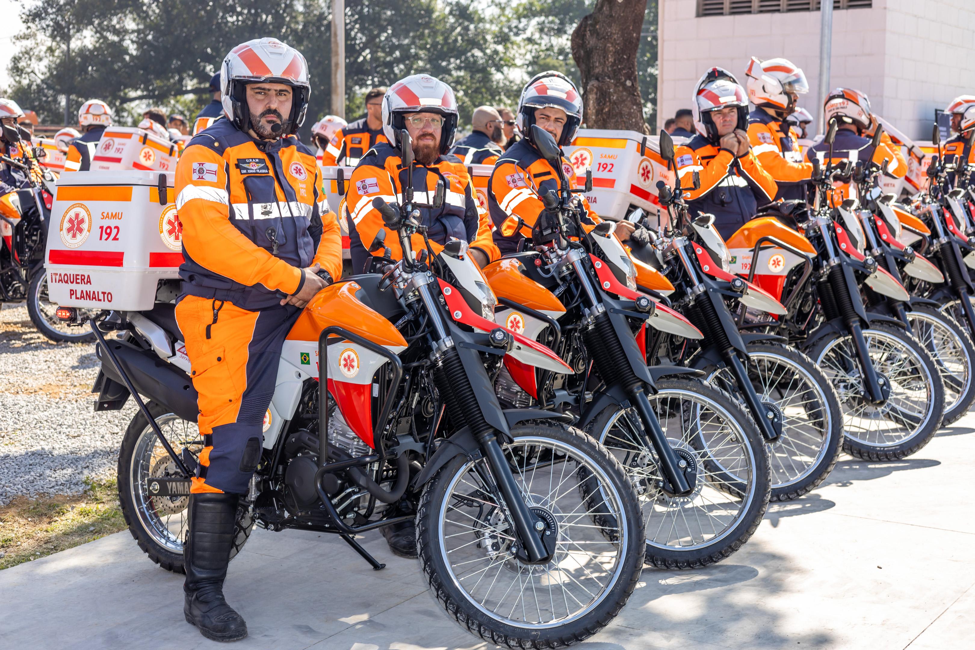 A imagem mostra um grupo de profissionais do SAMU (Serviço de Atendimento Móvel de Urgência) sentados em motos perfiladas, prontos para atuação emergencial. Todos vestem uniformes nas cores laranja vibrante, azul escuro e detalhes em branco refletivos, que garantem alta visibilidade. Os capacetes são brancos, com faixas em laranja e azul. Em destaque, um profissional está sentado sobre a moto, com expressão séria. Ele tem barba curta e escura, pele clara, e está olhando diretamente para a câmera. Outro profissional logo atrás possui barba mais longa e óculos transparentes. Ao fundo, há mais motociclistas, todos com o mesmo padrão de uniforme, indicando organização e preparo para atendimento rápido. As motos são adaptadas especialmente para emergências, na cor predominantemente preta, com partes em laranja e vermelho, contendo baús identificados com o número