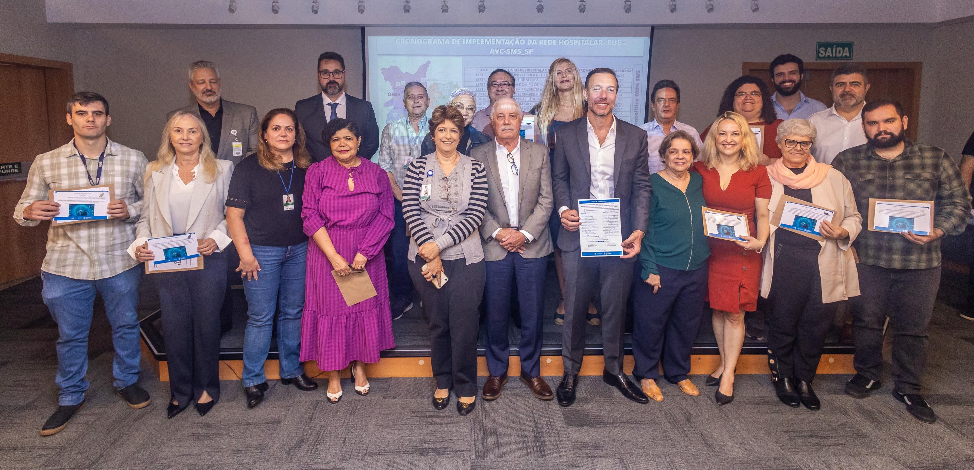 A imagem mostra um grupo de pessoas em um auditório, posicionadas para uma foto oficial. As pessoas estão distribuídas em duas fileiras. Na fileira frontal, todos estão em pé, alguns seguram certificados ou diplomas, indicando que participaram ou foram reconhecidos por algo específico no evento. Há uma diversidade aparente em termos de idade, estilo e trajes das pessoas presentes. À esquerda, há um homem jovem, pele clara, cabelo curto castanho escuro, vestindo uma camisa xadrez e calça jeans, segurando um certificado. Ao lado dele está uma mulher madura, pele clara, cabelo loiro liso médio, vestindo blazer branco e calça escura. Próximo, há uma mulher de cabelo castanho escuro e ondulado, pele clara, camiseta preta e jeans, usando crachá de identificação. Ao centro, uma mulher com vestido roxo vibrante, cabelo curto preto e pele morena clara, segura um envelope pardo nas mãos. Uma mulher ao lado dela usa blusa cinza listrada e colete cinza, tem cabelo castanho curto e pele clara. Um homem mais velho ao centro, pele clara, cabelo branco curto, usa terno cinza claro. Um homem mais alto e magro, pele clara, cabelo curto castanho claro, veste um terno azul escuro e camisa branca, segurando um documento. Há ainda uma mulher jovem, de vestido vermelho vibrante, cabelo loiro médio e pele clara, sorrindo enquanto segura um certificado. Outra mulher mais idosa, cabelo curto branco, pele clara, veste casaco claro e cachecol rosa claro. Na fileira de trás, há um grupo diverso de homens e mulheres com roupas mais formais, a maioria com trajes em tons escuros. Eles estão posicionados em um degrau mais elevado, o que possibilita a visibilidade de seus rostos.