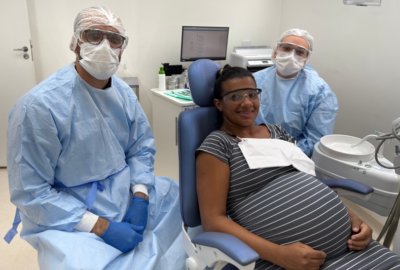 A imagem mostra uma mulher grávida sentada em uma cadeira de atendimento médico, sorrindo e parecendo feliz. Ela tem pele negra, cabelos escuros e curtos, e está vestindo uma blusa de riscas horizontais em tons de cinza e branco. Ela usa óculos de proteção e uma máscara facial, além de uma avental branco sobre a roupa. Na sua barriga, há uma toalha branca. Do lado esquerdo da imagem, há um profissional de saúde, que veste uma roupa de proteção azul, uma touca de plástico transparente, óculos de proteção e máscara facial. Esse profissional também usa luvas azuis e está de braços cruzados. Do lado direito, há outro profissional de saúde com roupas semelhantes, também usando touca, máscara, óculos de proteção, avental azul, e luvas, observando a cena. Ao fundo, há uma mesa com computador e outros equipamentos médicos, numa sala clara e bem iluminada.
