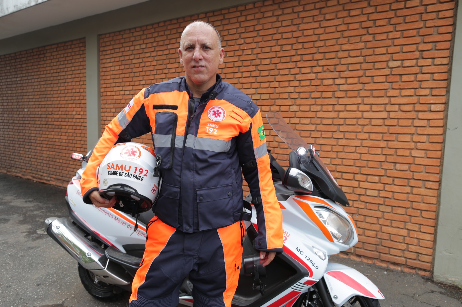 A imagem mostra Marcelo, um homem adulto, de pele clara e cabelos curtos e grisalhos, está em pé ao lado de uma motocicleta de emergência. Ele veste um uniforme de trabalho composto por uma jaqueta azul escura com detalhes em laranja neon e faixas reflexivas cinzas nos ombros e mangas, e calças compridas azuis escuras com detalhes laterais em laranja neon. Na parte superior da jaqueta, no lado direito do peito, há um patch com o texto