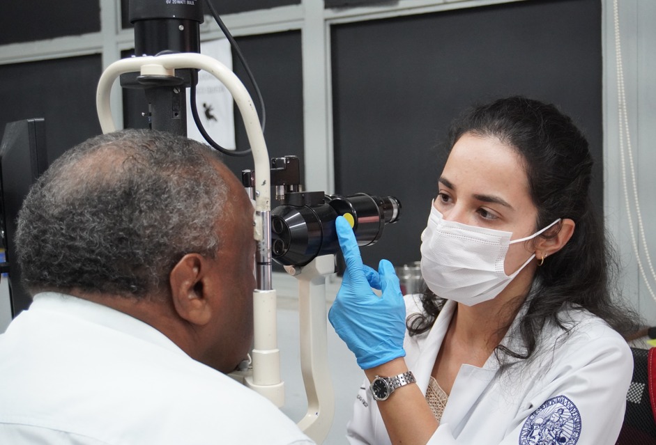 À direita, uma mulher de pele clara com cabelos longos, escuros e ondulados, presos parcialmente para trás, está inclinada sobre um equipamento médico. Ela usa uma máscara facial branca que cobre seu nariz e boca, e luvas azuis de procedimento em ambas as mãos. Em seu pulso esquerdo, ela usa um relógio de prata. Ela veste um jaleco branco com um brasão azul e branco na manga direita, que parece conter uma figura humana e texto ao redor. Seus olhos castanhos estão focados no paciente. Ela parece estar tocando ou ajustando o equipamento com o dedo indicador da mão esquerda.  À esquerda, um homem negro com cabelos curtos e grisalhos está sentado em frente ao equipamento. Vemos principalmente a lateral e a parte de trás de sua cabeça. Ele veste uma camisa branca. Seu olho direito está posicionado em frente à lente do equipamento.  O equipamento oftalmológico tem várias lentes, suportes brancos e pretos, e uma fonte de luz direcionada para o olho do paciente. Ao fundo, há uma parede com painéis cinzas e uma faixa horizontal preta na parte superior.