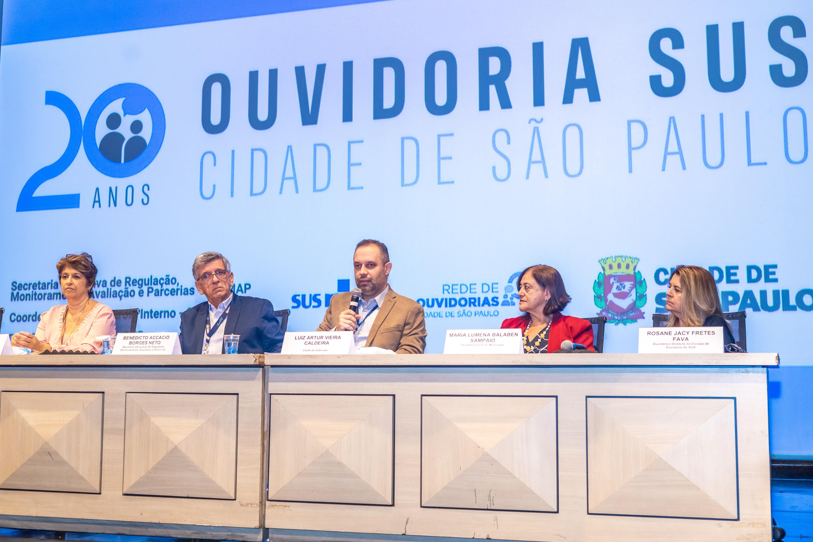 A imagem mostra uma mesa de conferência com cinco pessoas sentadas, participando de um evento em comemoração aos 20 anos da Ouvidoria SUS da Cidade de São Paulo. Ao fundo, há um painel com a identidade visual do evento e logotipos da Cidade de São Paulo, SUS, Secretaria Municipal da Saúde (SMS-SP), entre outros. Da esquerda para a direita, os participantes são: Uma mulher de roupa clara; Benedicto Accacio Borges Neto (identificado pela placa), provavelmente representando um órgão de controle interno; Luiz Artur Vieira Caldeira, que segura um microfone e parece estar falando – identificado como chefe de gabinete da Secretaria Municipal da Saúde; Maria Lumena Balaben Sampaio, também identificada por uma placa; Rosane Jacy Fretes Fava, da Ouvidoria-Geral do SUS (Ministério da Saúde). O evento celebra e reforça a importância da ouvidoria como canal de escuta e participação cidadã no SUS paulistano.