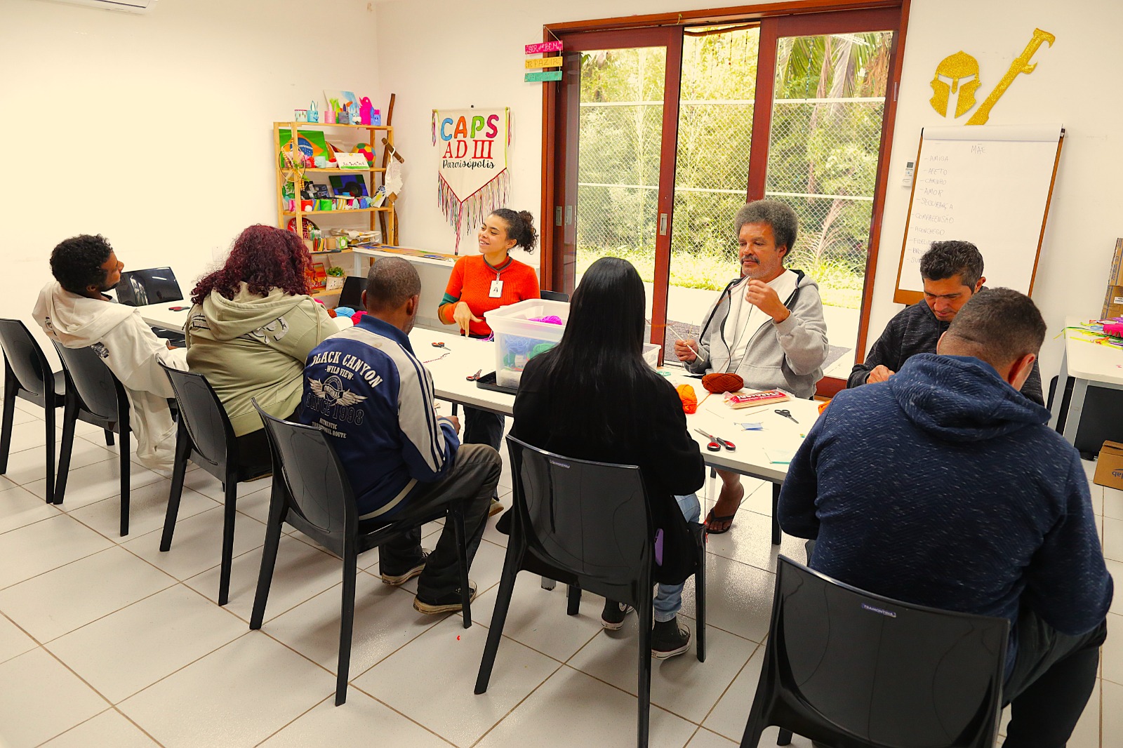 A imagem mostra um grupo de sete pessoas reunidas em torno de uma longa mesa branca em uma sala bem iluminada. Eles estão envolvidos numa atividade de artesanato. Começando pela esquerda, a primeira pessoa visível é um homem de pele negra, com cabelos curtos e escuros. Ele está de costas para a câmera e veste uma blusa branca com capuz. Ao lado dele, uma pessoa com pele em um tom mais claro e cabelos cacheados e volumosos, tingidos em tons de vermelho ou vinho, está sentada de costas para a câmera. Veste um casaco bege claro com capuz. A terceira pessoa a partir da esquerda é um homem de pele negra, com cabelos curtos e escuros. Ele está de perfil, olhando para a direita, e veste uma jaqueta azul escura com mangas brancas. Na cabeceira da mesa, no lado esquerdo, está uma mulher com cabelos escuros presos em um rabo de cavalo baixo. Ela veste uma blusa laranja e parece estar falando ou interagindo com o grupo. Atrás dela, na parede, há uma decoração colorida com o texto
