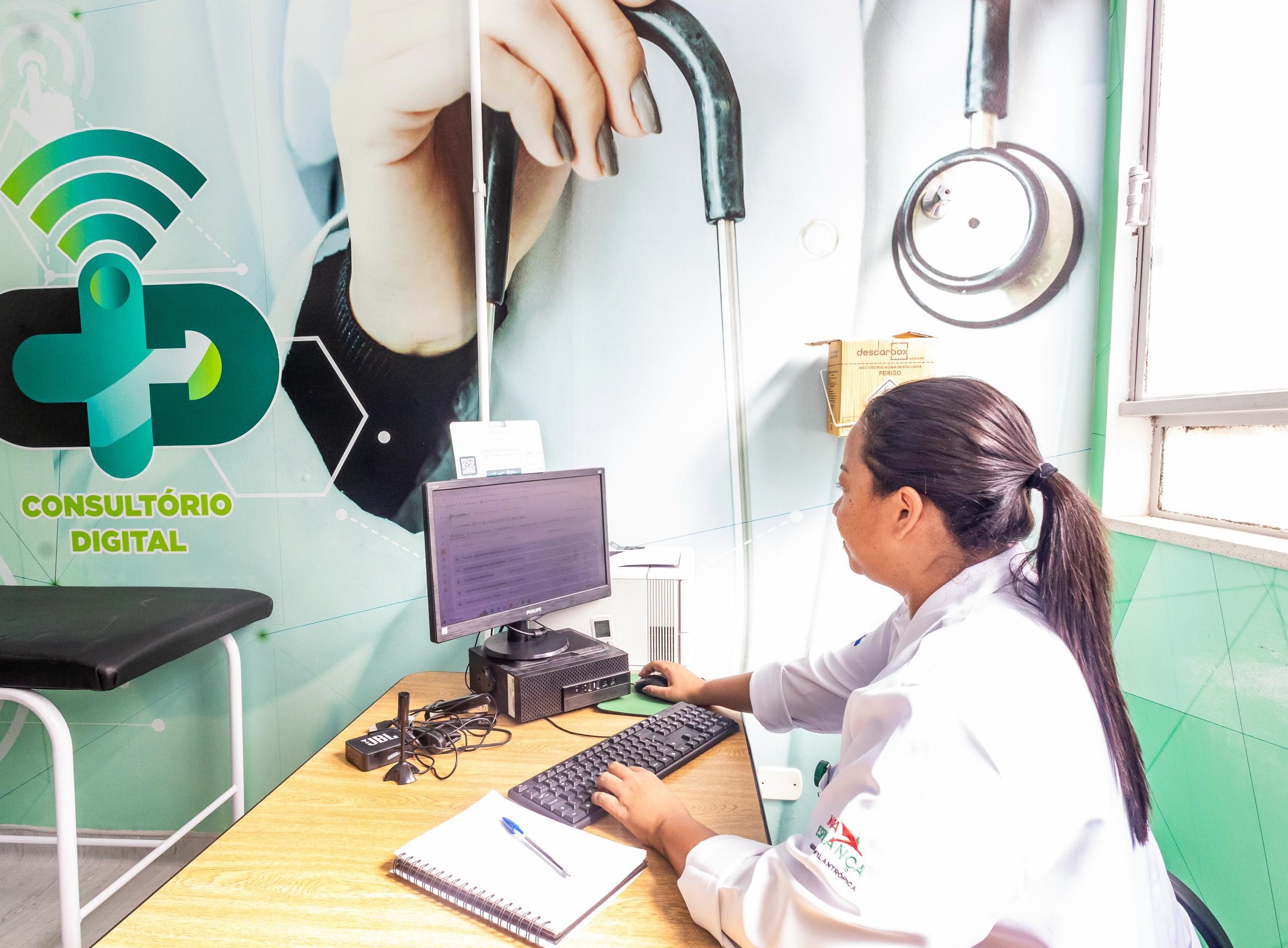 A imagem mostra uma profissional da saúde trabalhando em um consultório digital. Ela está sentada à mesa, utilizando um computador de mesa. Na parede, há um grande adesivo com o logotipo do Consultório Digital, destacando um símbolo de saúde A profissional usa jaleco branco, com um logotipo bordado na manga. O ambiente é bem iluminado, organizado e demonstra uma estrutura voltada para atendimentos médicos remotos ou informatizados, representando a modernização do serviço público de saúde