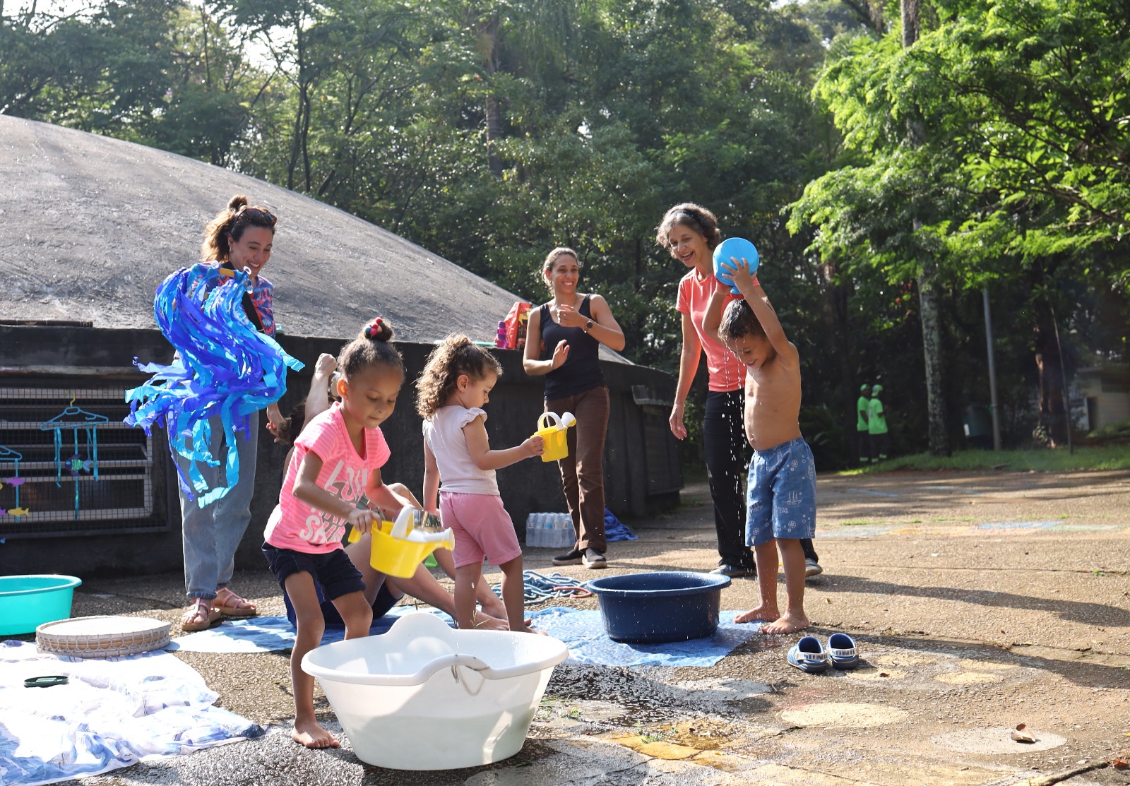 A imagem captura um momento de brincadeira ao ar livre com várias pessoas e crianças em um dia ensolarado. À esquerda, uma mulher adulta de pele clara e cabelos castanhos presos em um coque está envolvida em uma brincadeira com fitas azuis brilhantes, que parecem estar presas a um objeto não totalmente visível. Ela veste uma camiseta estampada e calças jeans claras, além de sandálias marrons. Ao lado dela, uma menina de pele clara com cabelos escuros cacheados, presos em dois pequenos coques no topo da cabeça, está brincando com um balde branco. Ela veste uma camiseta rosa com desenhos e shorts azuis escuros. No centro, outra menina de pele clara com cabelos castanhos cacheados, soltos e com uma presilha rosa, está segurando um regador amarelo. Ela veste uma camiseta branca e shorts rosa. Ao fundo, à esquerda, uma mulher adulta de pele clara com cabelos castanhos presos está sorrindo e olhando na direção das crianças. Ela veste uma regata preta e calças marrons. À direita, uma mulher adulta de pele clara com cabelos castanhos soltos está sorrindo e ajudando um menino. O menino, de pele clara e cabelos curtos e escuros, está segurando uma bola azul acima da cabeça. Ele está sem camisa e veste shorts azuis estampados. No chão, há uma lona azul escura com respingos de água, indicando que estão brincando com água. Há também um balde azul escuro, um balde branco maior com água, uma bacia azul clara e uma esteira de palha redonda. Um par de chinelos azuis está no chão, perto do menino e da mulher à direita. O cenário ao fundo é um espaço aberto com árvores altas e folhagem verde, sugerindo um parque ou área natural. A luz do sol projeta sombras no chão, indicando que é durante o dia. Uma estrutura com um telhado curvo e janelas gradeadas é visível ao fundo, à esquerda. As pessoas parecem estar relaxadas e se divertindo juntas.