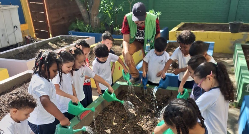 A imagem mostra uma atividade de educação ambiental com crianças em uma horta escolar. Um grupo de aproximadamente doze crianças, vestindo uniformes escolares, está reunido em torno de canteiros elevados de terra. Todas estão segurando regadores verdes e participando do momento de rega das plantas, enquanto um educador ou monitor, de boné e colete verde, as orienta.  O ambiente é ao ar livre, cercado por muros pintados com cores vivas e áreas de plantio bem organizadas. A interação é visivelmente alegre e colaborativa, com as crianças concentradas na tarefa e o adulto incentivando a participação.