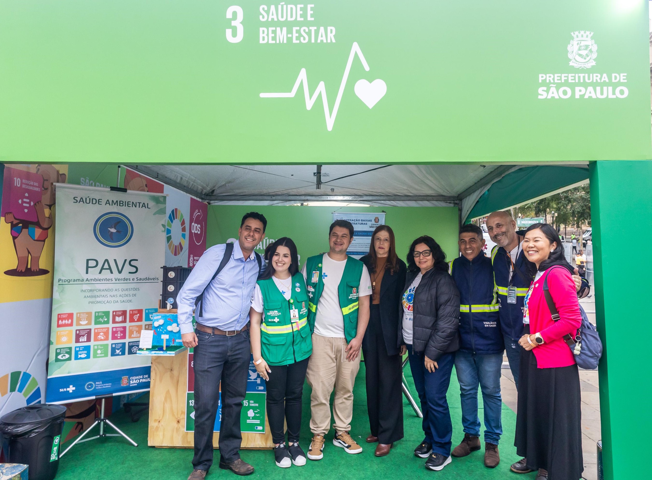A imagem mostra um grupo de nove pessoas posando sorridentes em frente a uma tenda verde identificada com o símbolo do Objetivo de Desenvolvimento Sustentável (ODS) 3 – Saúde e Bem-Estar, da Prefeitura de São Paulo. A tenda abriga o estande do Programa Ambientes Verdes e Saudáveis (PAVS), ligado à Saúde Ambiental, como indica o banner exposto no local. Os integrantes do grupo estão vestidos com diferentes trajes: alguns com coletes verdes do programa, outros com uniformes azuis da vigilância sanitária e trajes casuais. A ambientação inclui cartazes coloridos com os ícones dos ODS, destacando metas como saúde, educação, igualdade de gênero, cidades sustentáveis, entre outros. O cenário urbano e a arquitetura ao fundo sugerem que o evento ocorre em um espaço público da cidade.