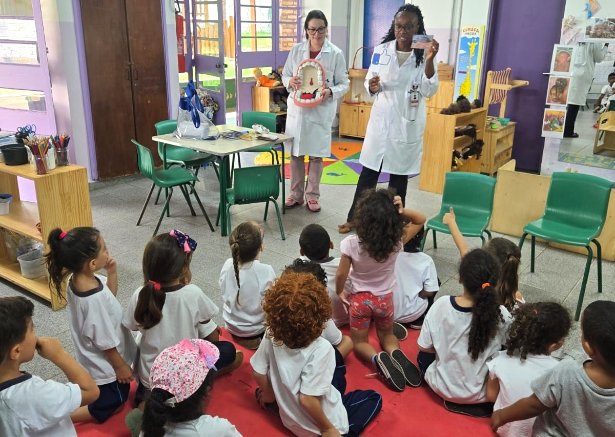 A imagem mostra uma atividade educativa sobre saúde bucal em uma sala de aula infantil. Duas profissionais da área da saúde, vestindo jalecos brancos, estão de pé à frente da sala conduzindo a apresentação. Uma delas segura um modelo gigante de boca com dentes, provavelmente para demonstrar técnicas de escovação. A outra exibe um cartaz com um símbolo de