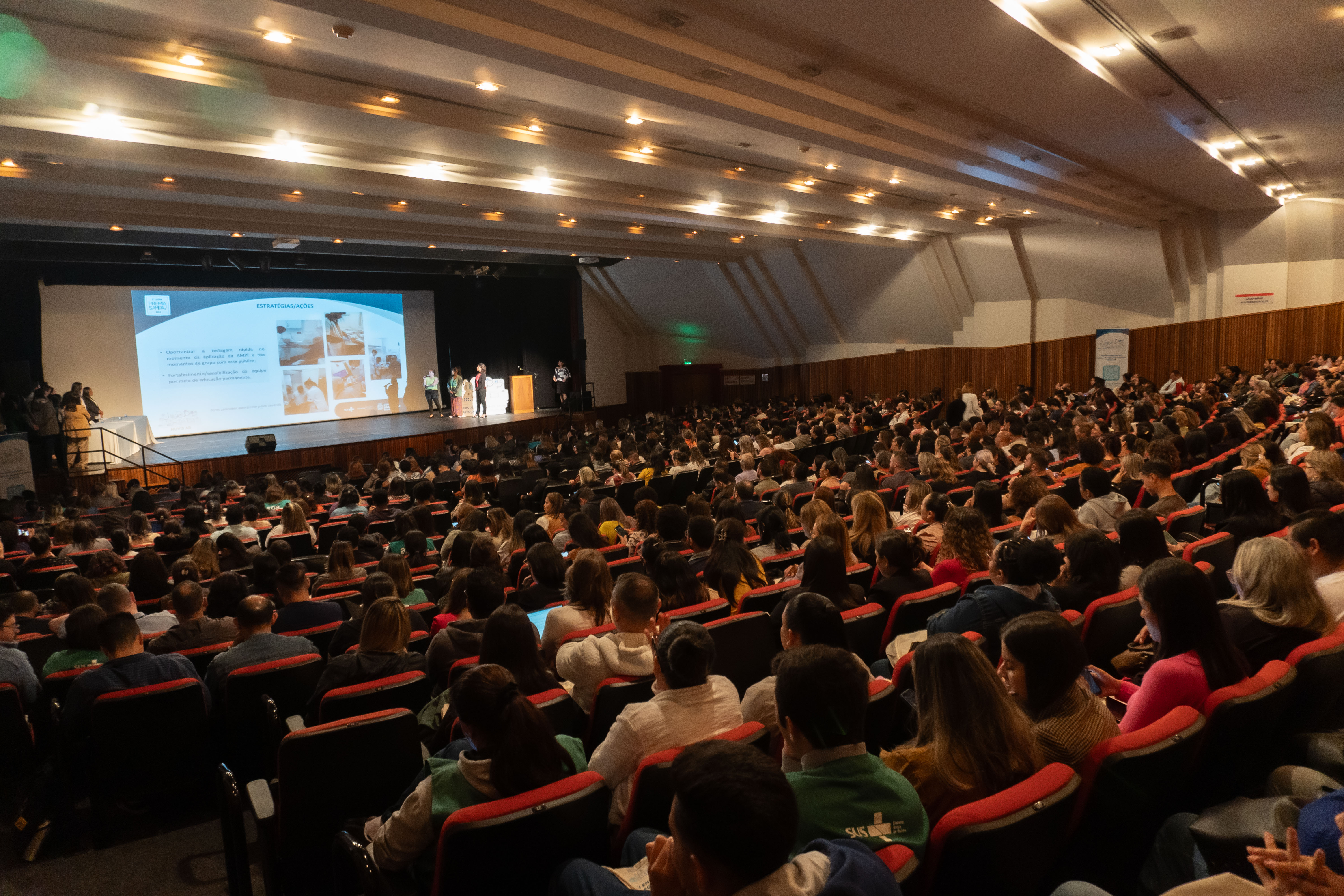 A imagem mostra um auditório amplo, lotado de pessoas assistindo a uma apresentação. O palco ao fundo possui um telão projetando slides, e algumas pessoas estão em pé fazendo uma apresentação. 