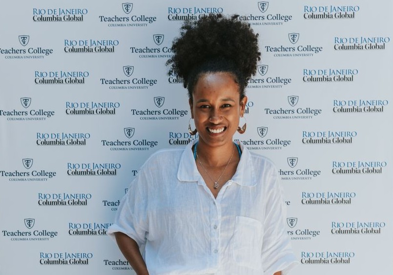A imagem mostra Fernanda, uma mulher negra, sorridente posando em frente a um painel de fundo branco com logos e textos de