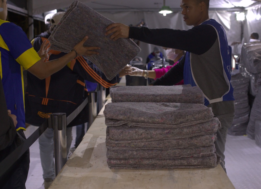 A foto mostra uma ação de distribuição de cobertores. Em primeiro plano, há uma pilha de cobertores dobrados sobre uma mesa comprida. Um homem usando colete azul — característico de voluntários ou agentes públicos — entrega um cobertor a uma pessoa do outro lado da mesa. A imagem está em um ambiente coberto, com iluminação artificial e outras pessoas ao fundo também participando da entrega ou recebimento de cobertores. A cena sugere um esforço organizado de acolhimento, especialmente em contexto de baixas temperaturas ou vulnerabilidade social.