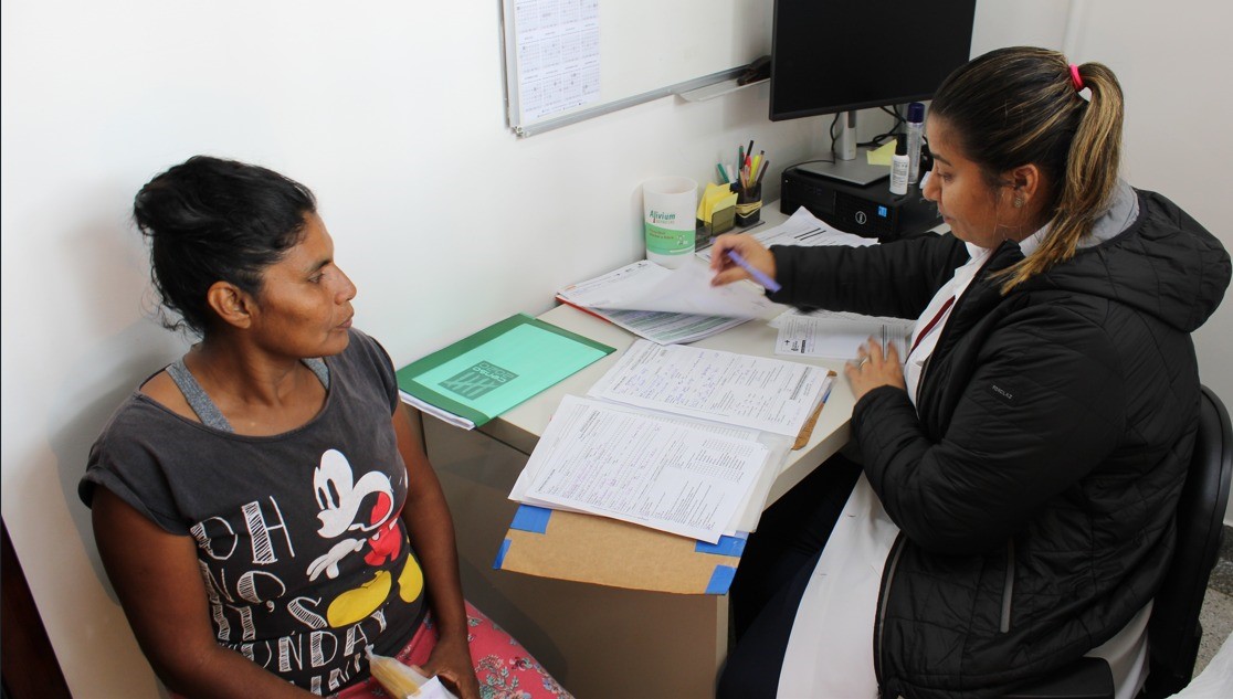 Da esquerda para direita, uma mulher indígena está sentada na cadeira do consultório e está de frente para a médica que está observando as fichas de saúde da paciente.  A paciente indígena está vestida com camiseta preta, com estampa do Mickey Mouse, e saia vermelha. A médica, que está sentada do lado direito, está observando as fichas e segurando uma caneta lilás. A médica tem os cabelos pretos e mechas loiras. Além do jaleco branco, ela está vestida com uma jaqueta na cor preta.