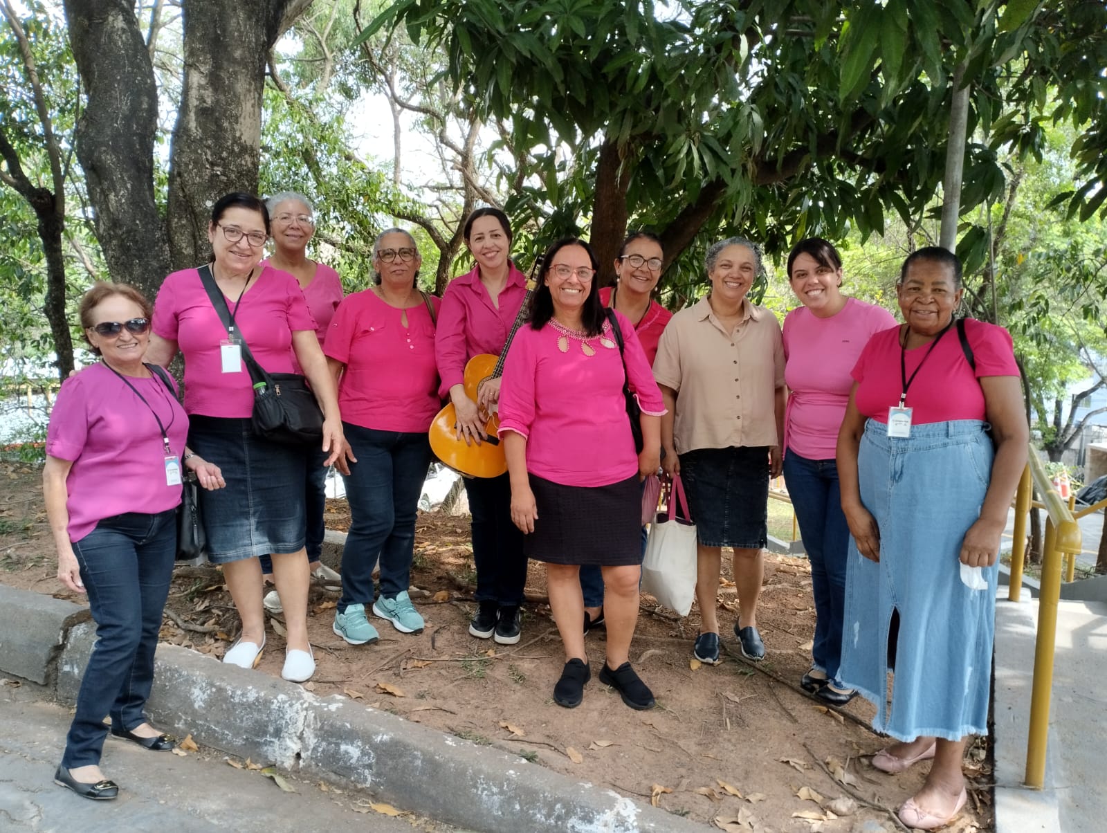 A imagem mostra um grupo de 11 mulheres reunidas em um ambiente externo, sob árvores. Dez delas vestem roupas em diferentes tons de rosa. Uma delas segura um violão.