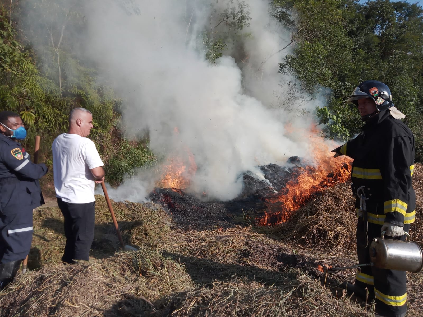 Na imagem, vemos uma simulação de combate a incêndio realizada durante um curso de formação de brigadistas, em uma área de vegetação, onde um foco de fogo controlado consome material orgânico seco enquanto libera densa fumaça branca. Três participantes — dois deles usando equipamentos de segurança, incluindo uniforme e capacete — observam e atuam na contenção das chamas com o uso de abafadores e técnicas manuais. O cenário revela a importância do treinamento prático para atuação rápida e eficaz em situações reais de queimadas, especialmente em regiões de mata.
