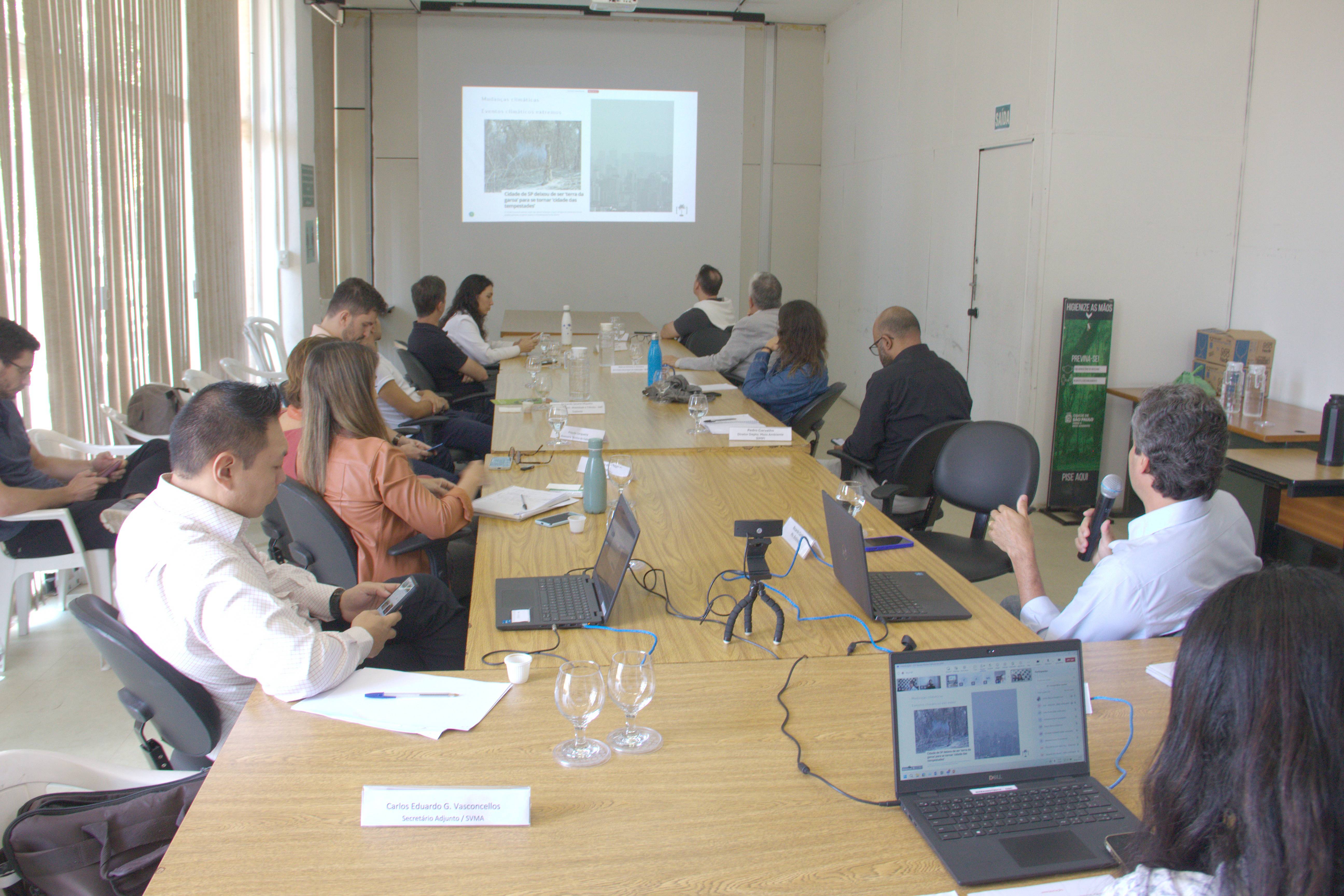 Reunião do Conselho Municipal do Meio Ambiente e Desenvolvimento Sustentável (CADES), com participantes reunidos ao redor de uma mesa retangular em uma sala da Secretaria do Verde e do Meio Ambiente (SVMA). Um dos conselheiros conduz a apresentação, enquanto um projetor exibe slides com imagens de áreas de vegetação