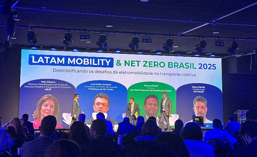 Painel do evento Latam Mobility & Net Zero Brasil 2025, com o tema “Desmistificando os desafios da eletromobilidade no transporte coletivo”. A apresentação ocorreu em um palco com iluminação em tons de azul, roxo e verde, cores predominantes também no telão de fundo, que destaca os nomes e cargos dos palestrantes. Entre os participantes, está o secretário municipal de Transportes e Mobilidade Urbana de São Paulo, Luiz Felipe Aryalano.