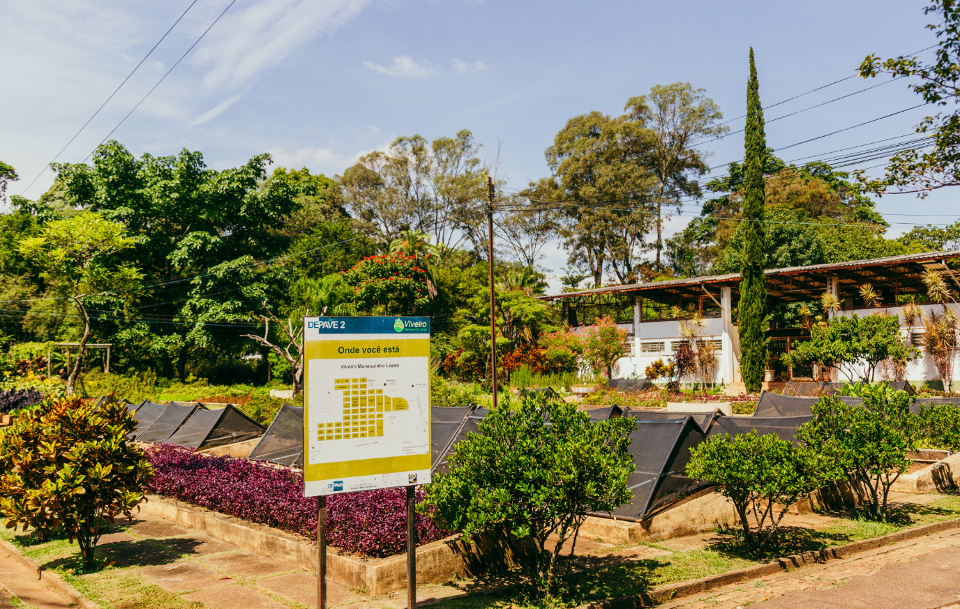 A imagem mostra uma área do Viveiro Manequinho Lopes, com canteiros de cultivo organizados em fileiras protegidas por telas sombreadoras, rodeados por uma vegetação rica e diversa composta por arbustos, árvores e plantas ornamentais. Em primeiro plano, há uma placa com o título